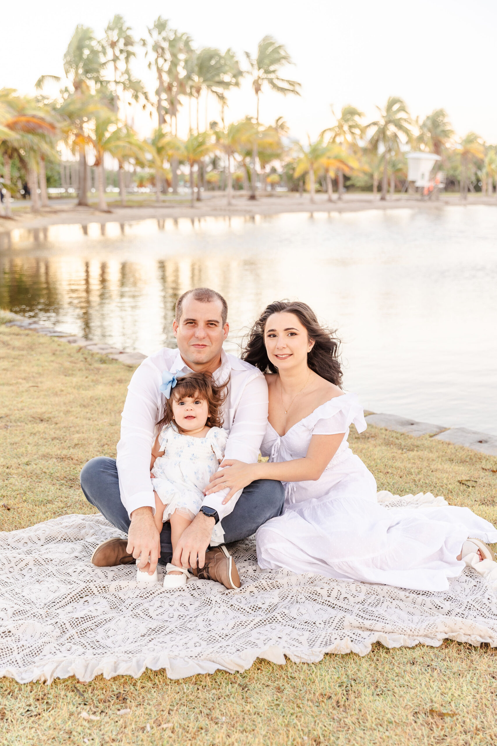 A smiling toddler girl in a white dress sits in dad's lap on a lace picnic blanket in a waterfront park with mom sitting beside them before visiting water parks near fort lauderdale