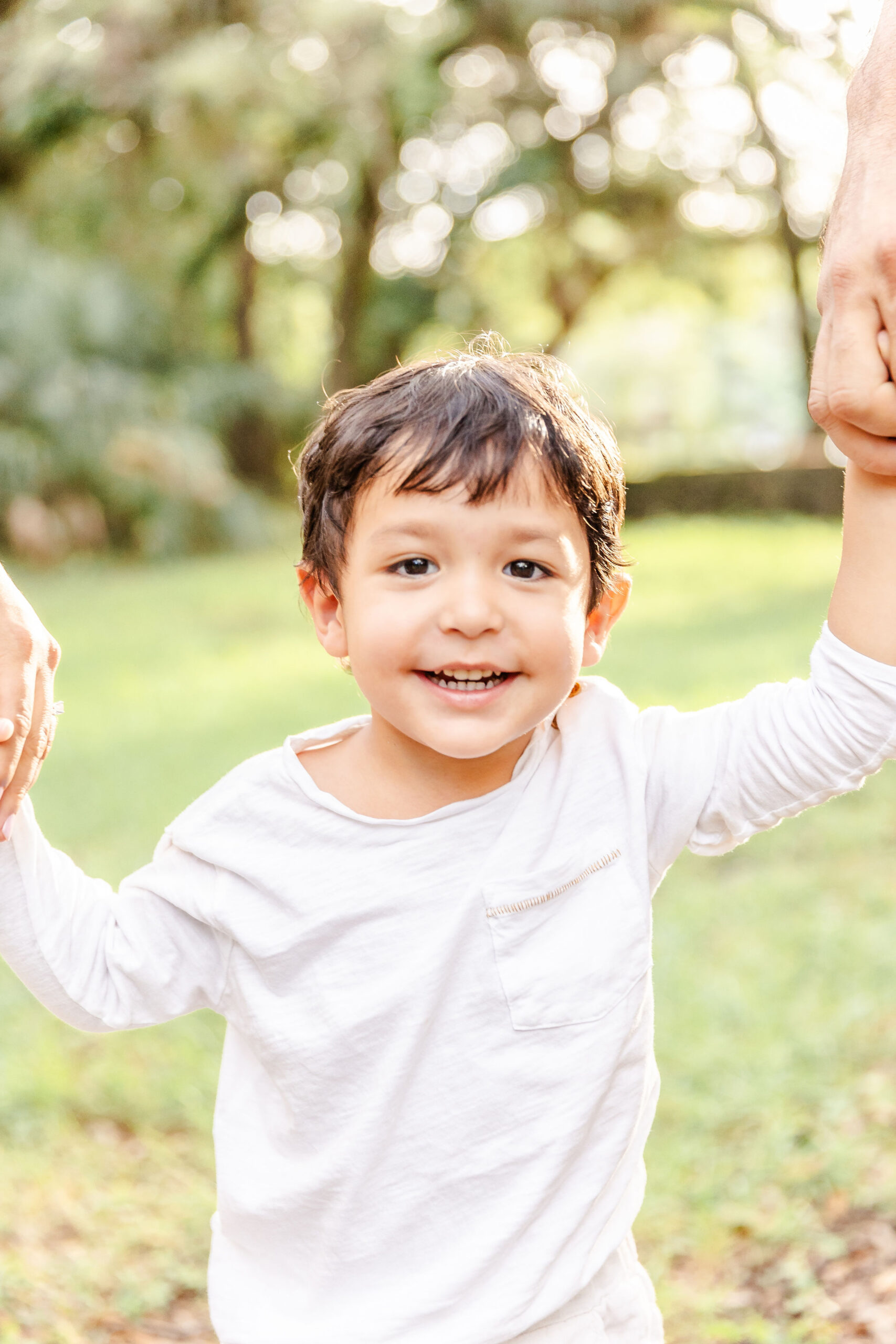 A smiling toddler boy holding mom and dad's hands in a park at sunset in a white shirt