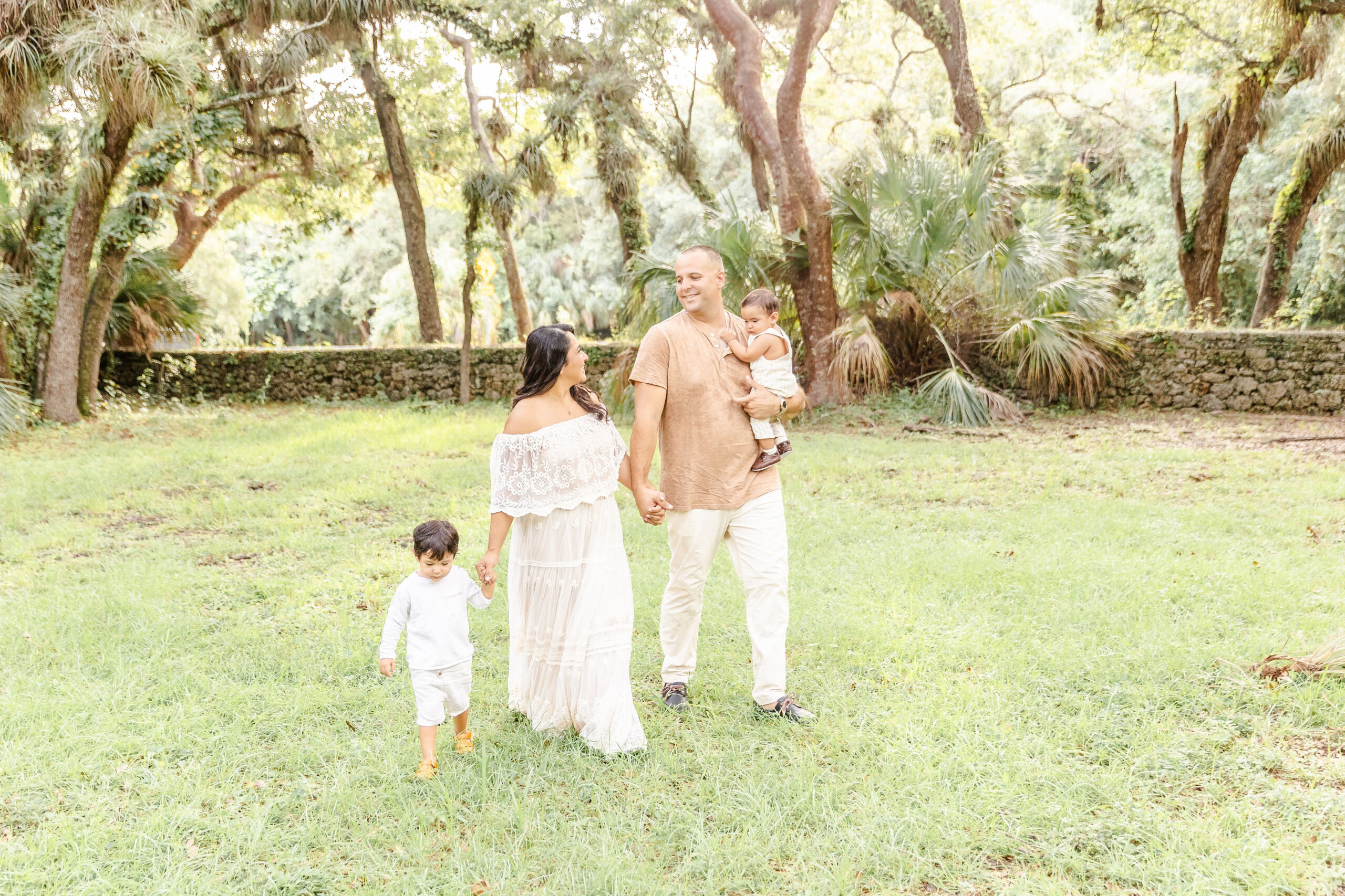 A mom and dad walk holding hands and smiling while holding hands with their toddler son and an infant on dad's arm during fall activities in fort lauderdale