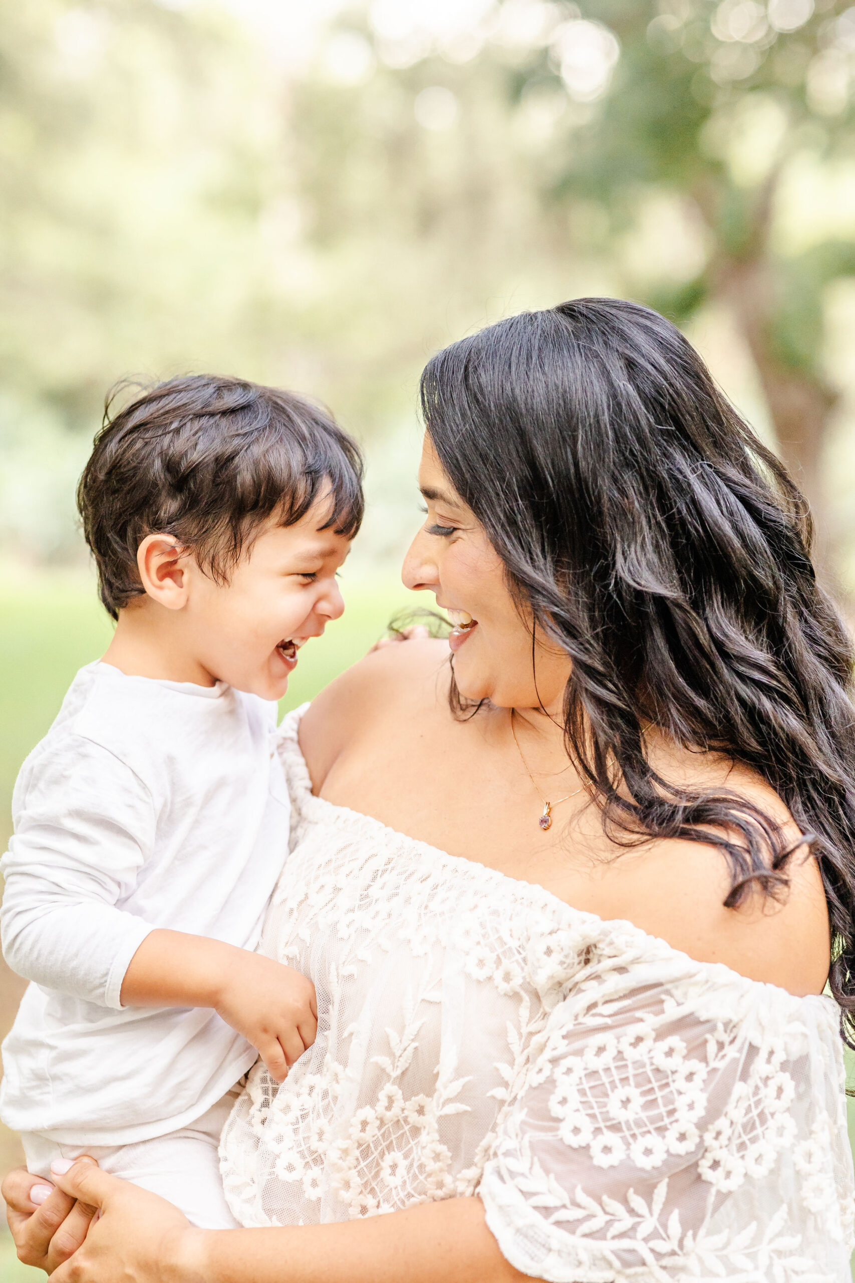 A toddler in white giggles while playing on mom's hip in a park in a lace dress before some fall activities in fort lauderdale