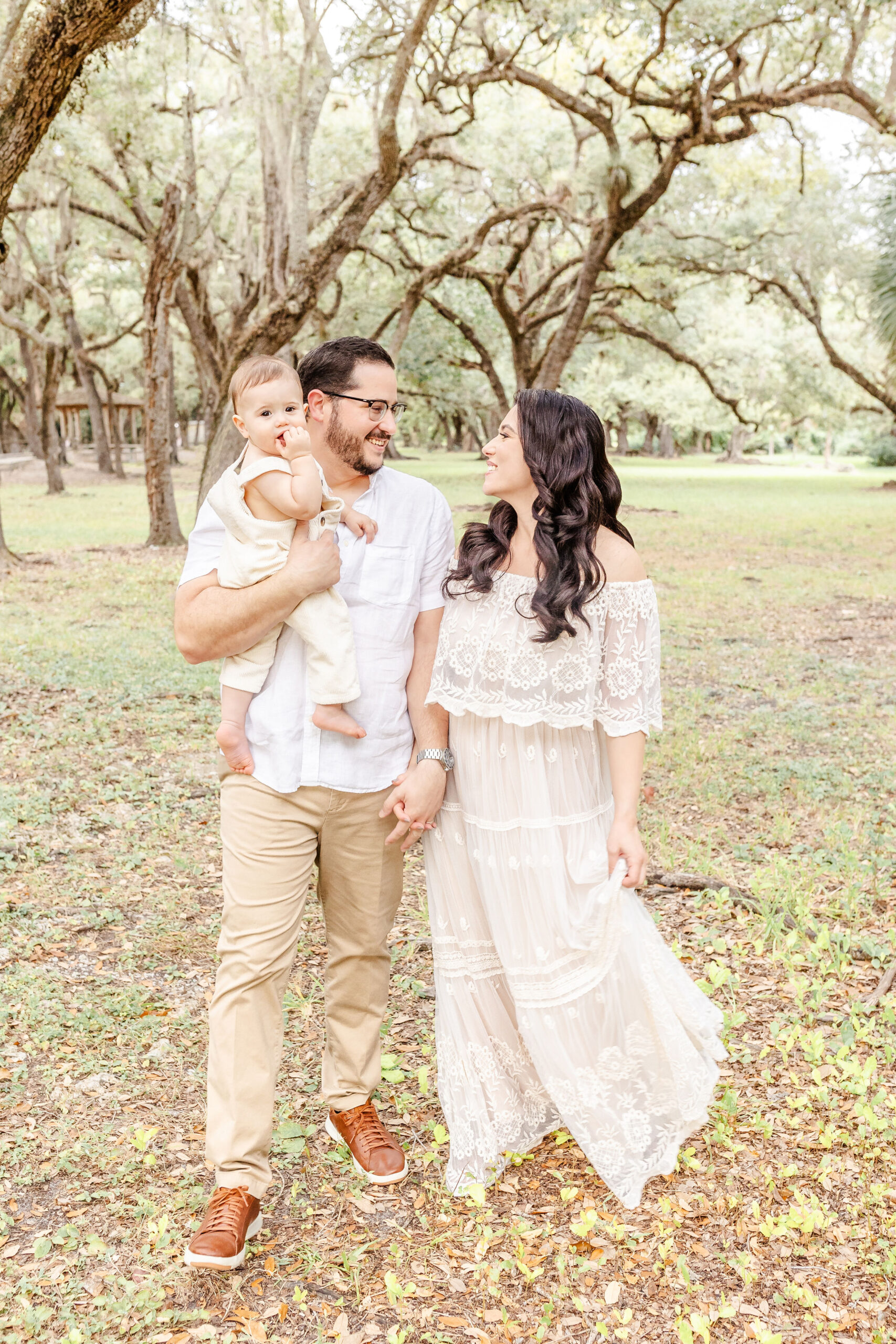 A mom and dad in white walk holding hands in a Florida park while dad holds their infant