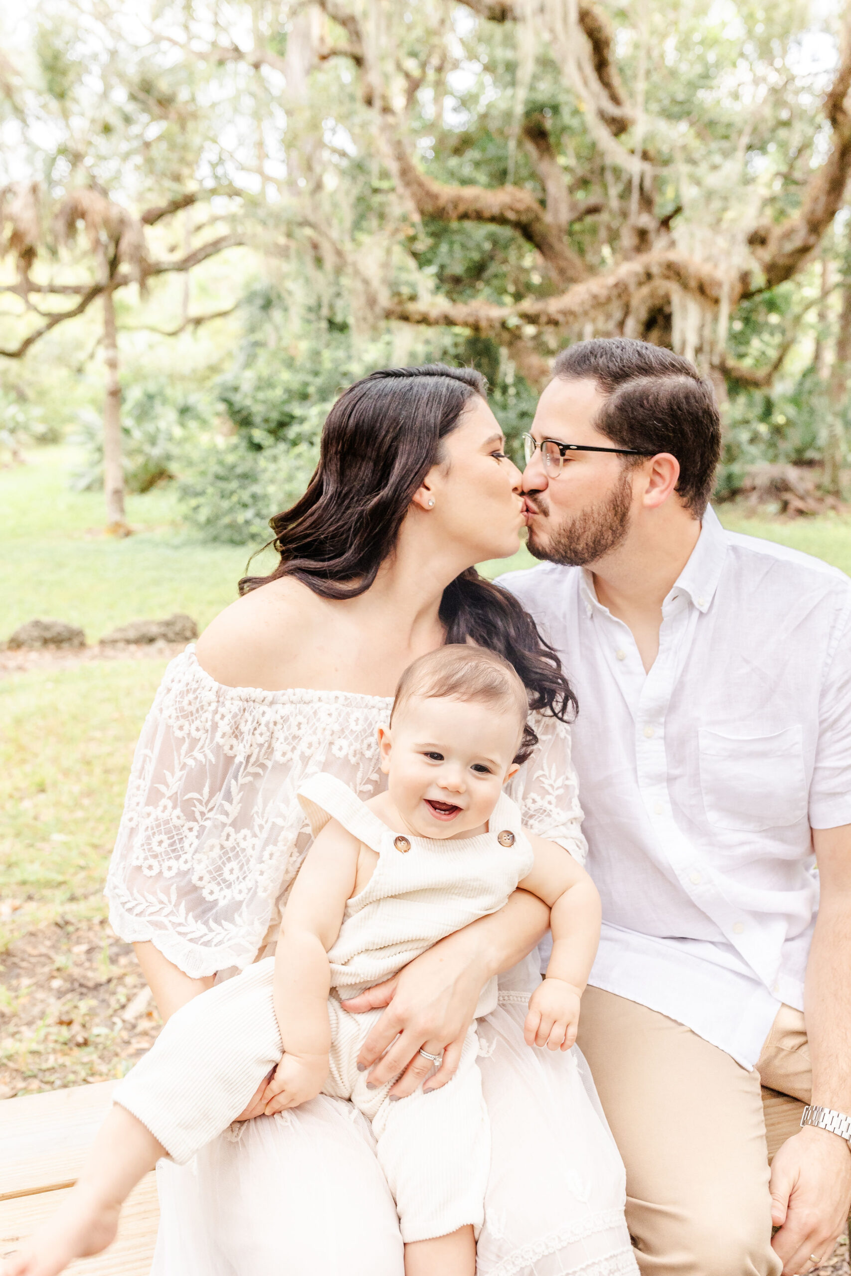 Mom and dad in white kiss while their happy infant sits in mom's lap in white overalls on a park bench before visiting a pumpkin patch in fort lauderdale