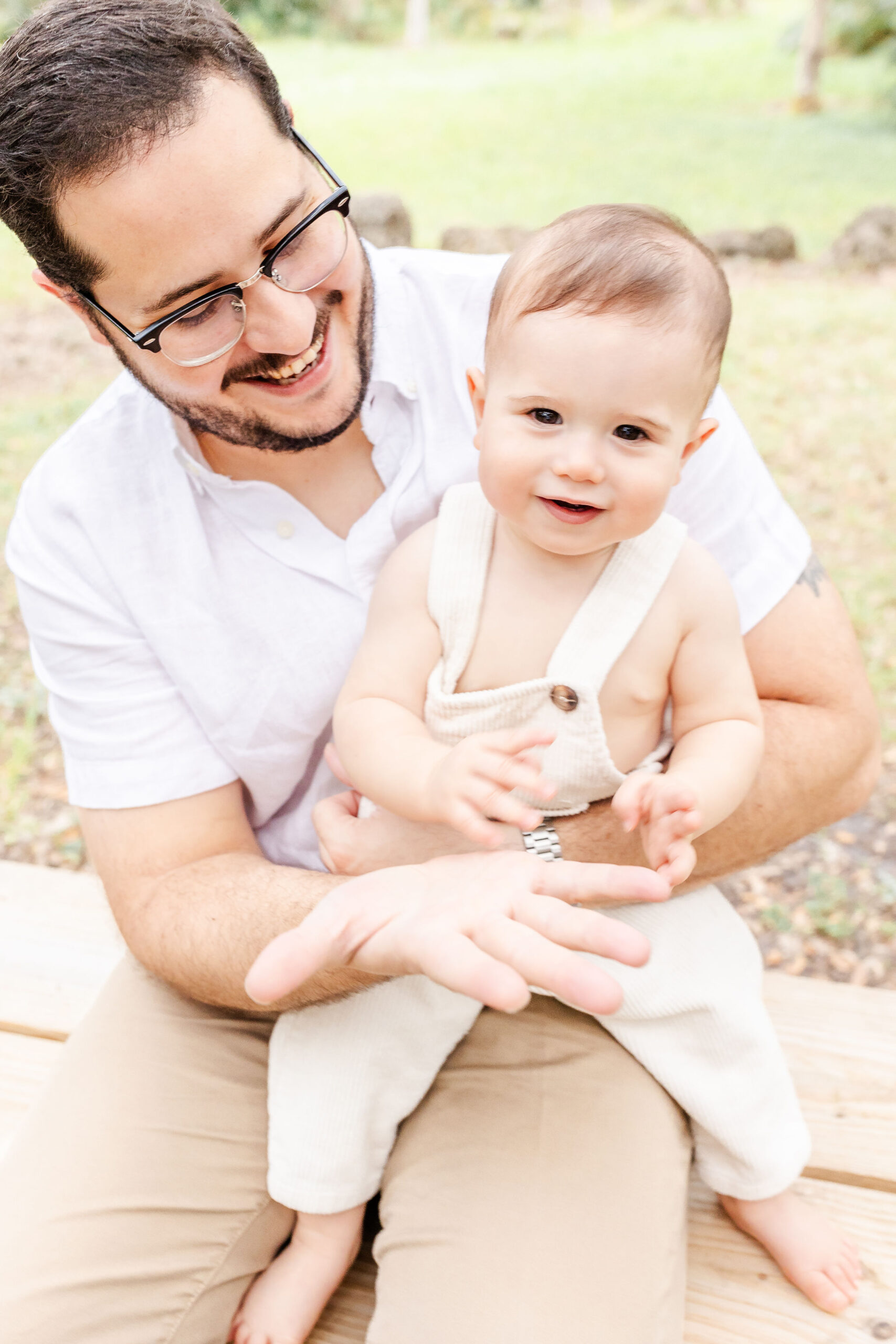 A happy infant in cream overalls plays in dad's lap on a park bench before visiting a pumpkin patch in fort lauderdale