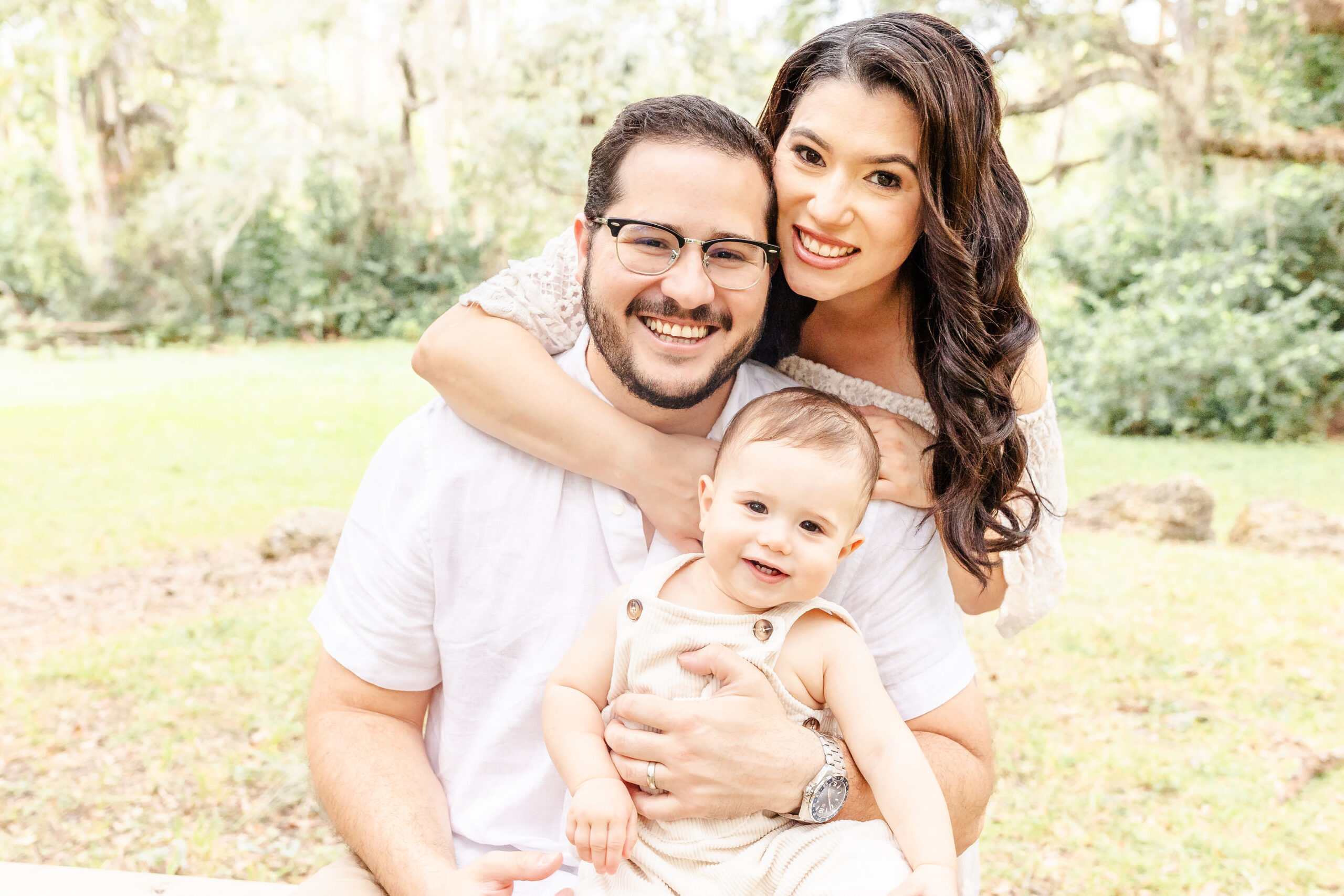 A happy mom and dad snuggle in a park in white witht heir infant in dad's lap