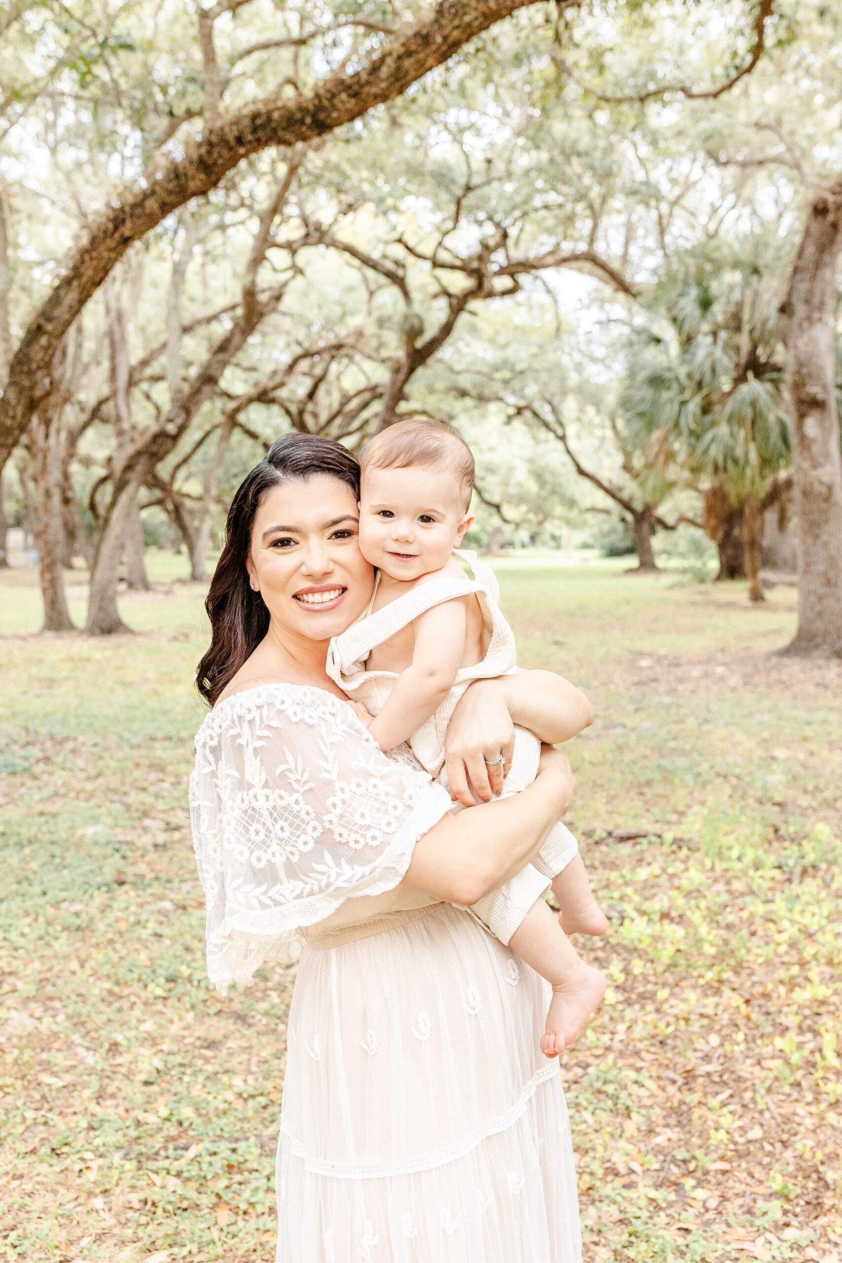 A happy mom in a white dress stands hugging her infant son in white overalls in a park at sunset before enjoying christmas lights in fort lauderdale