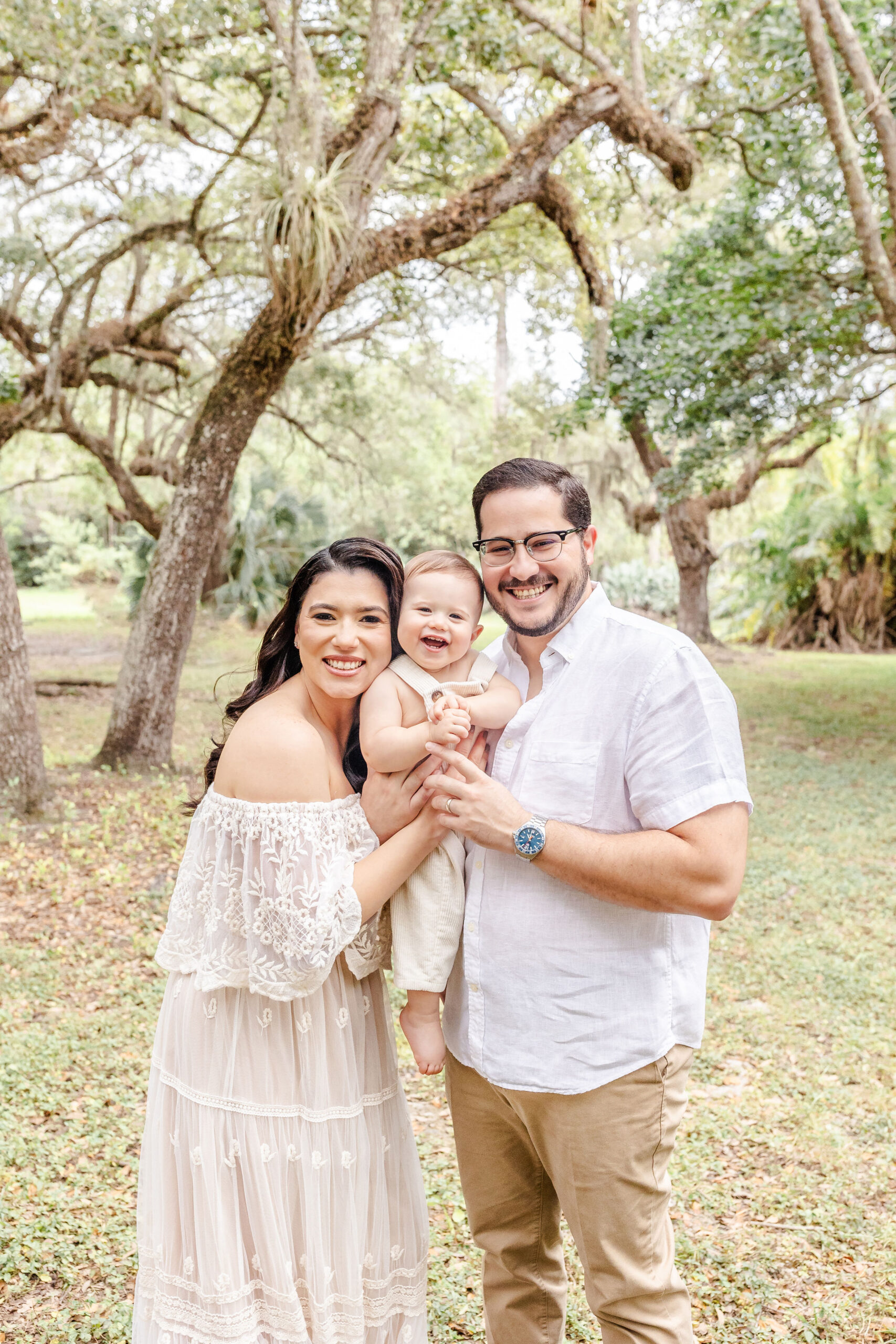 A giggling infant is held between mom and dad in a park at sunset all dressed in white before visiting christmas lights in fort lauderdale