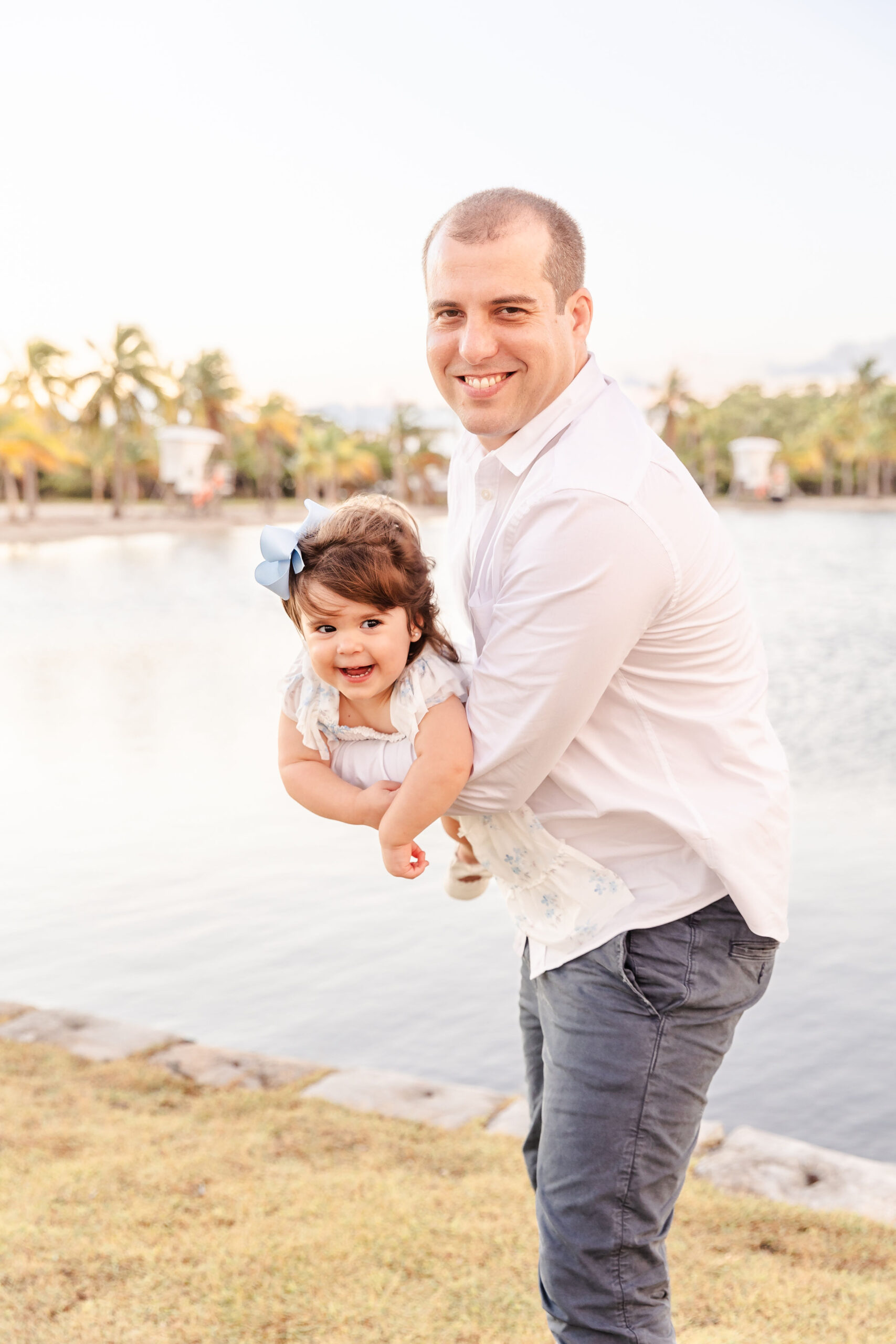 A happy dad in a white shirt helps his daughter fly in a park lawn by the water before exploring christmas trees in fort lauderdale