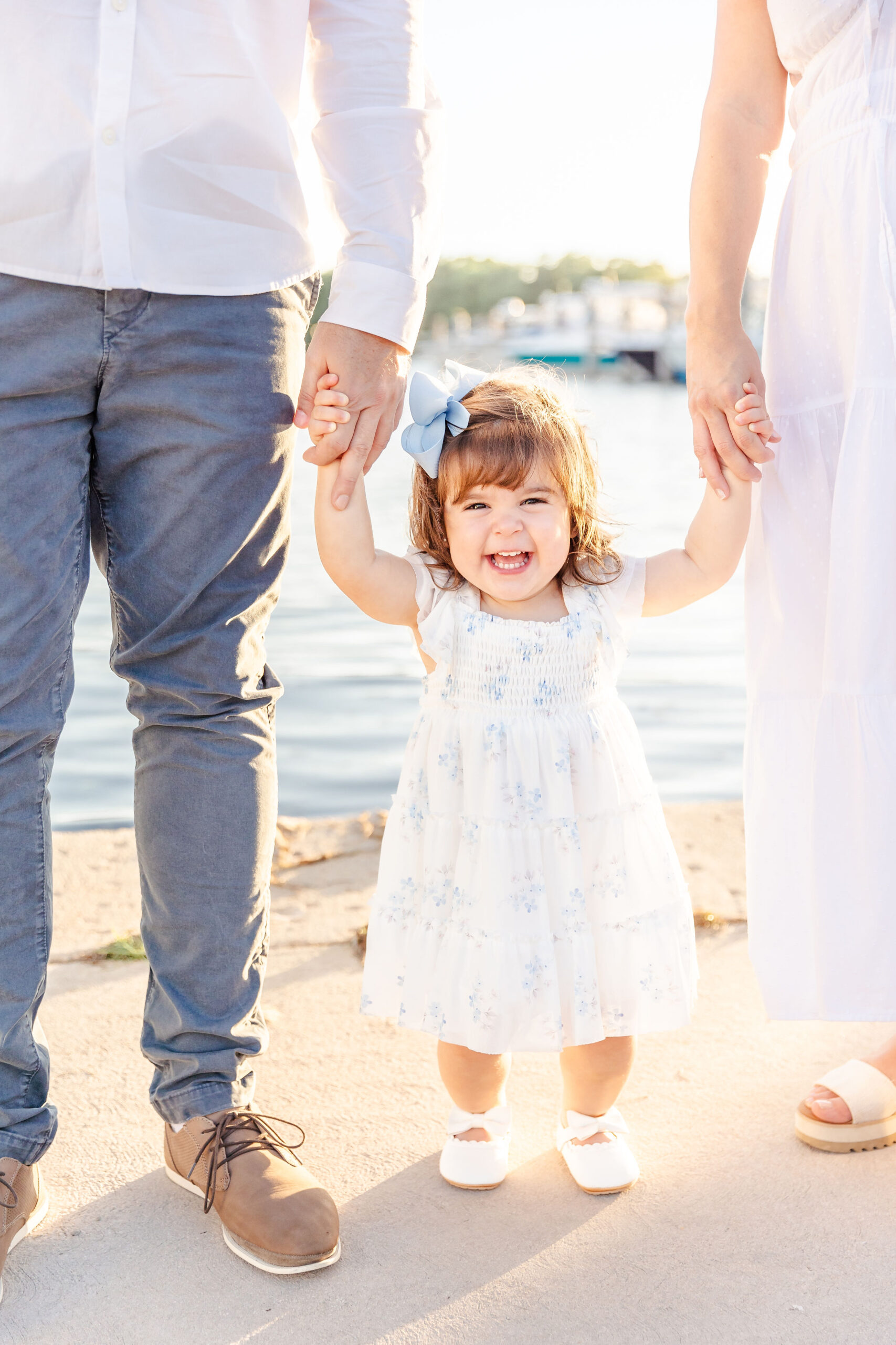 A giggling toddler girl stands by the water holding mom and dad's hands in a white dress before finding christmas trees in fort lauderdale