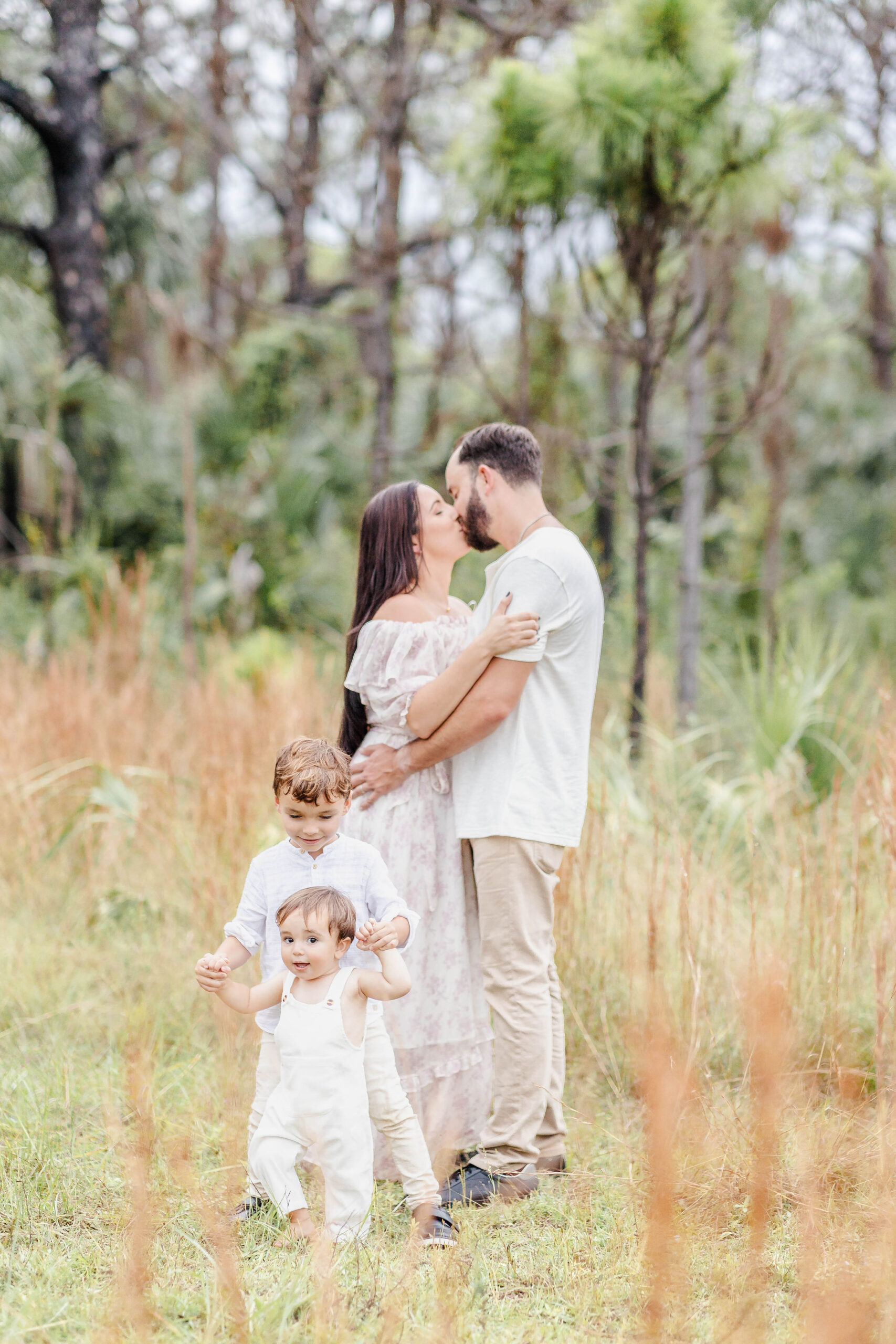 Mom and dad wearing white kiss as their two toddler sons play at their feet in a field of tall grass