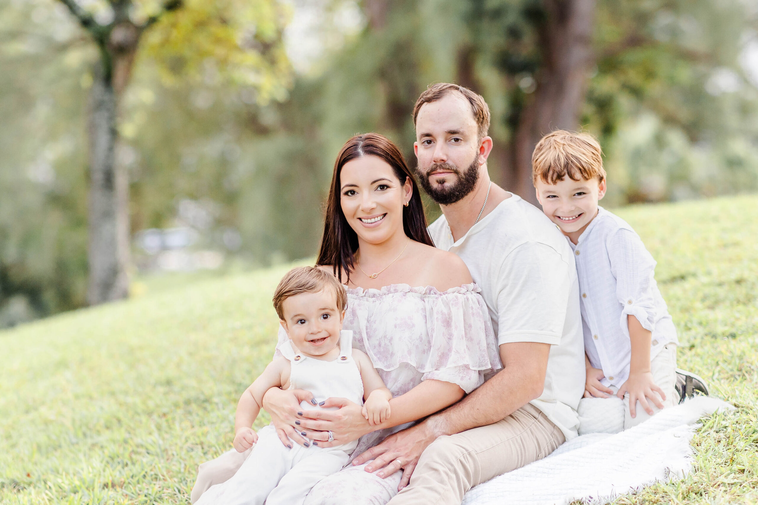A mom and dad sit on a picnic blanket in a park lawn witht heir two toddler sons all in white before finding christmas trees in miami