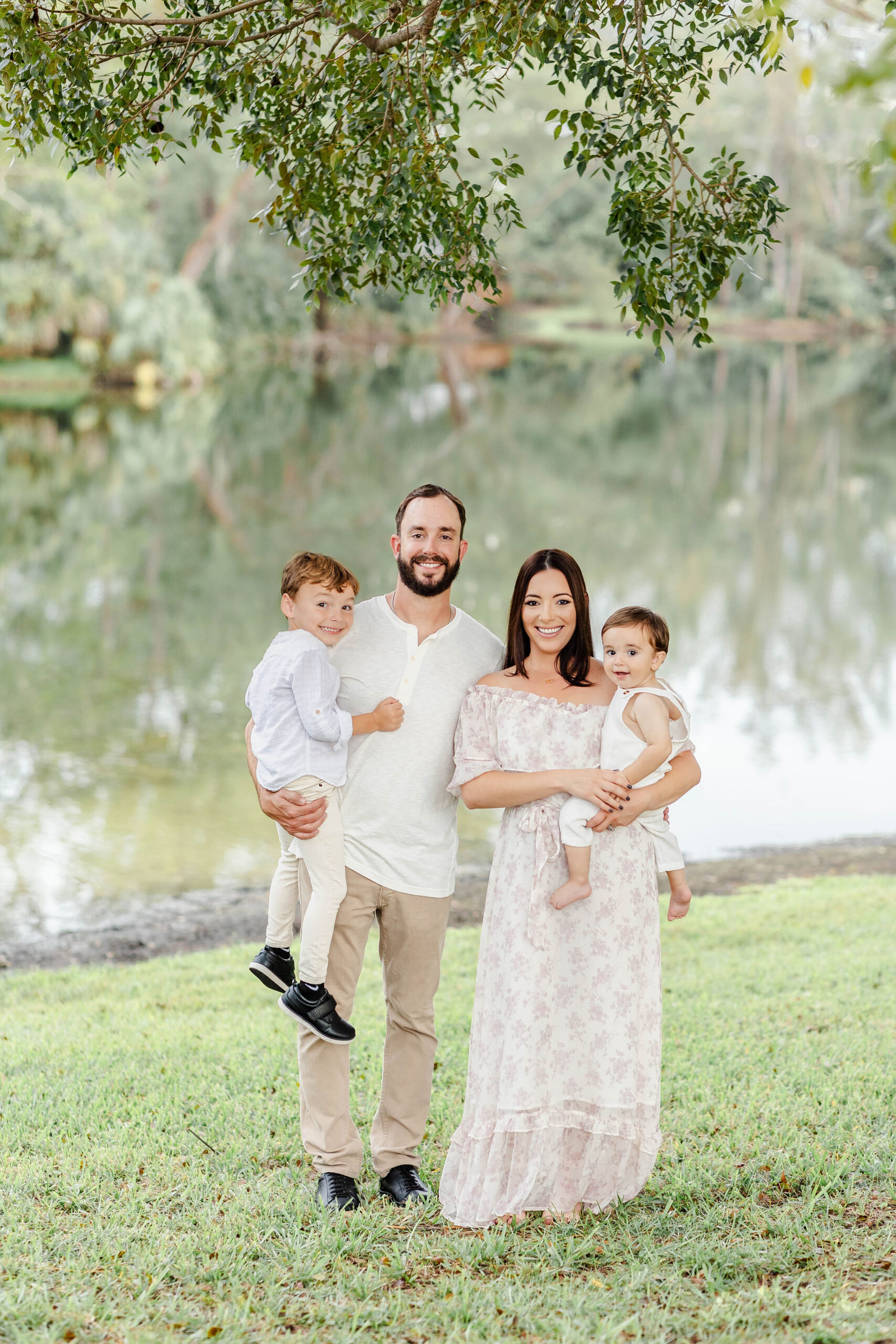 A mom and dad hold their two toddler sons on their hips by a lake in a park before finding christmas trees in miami