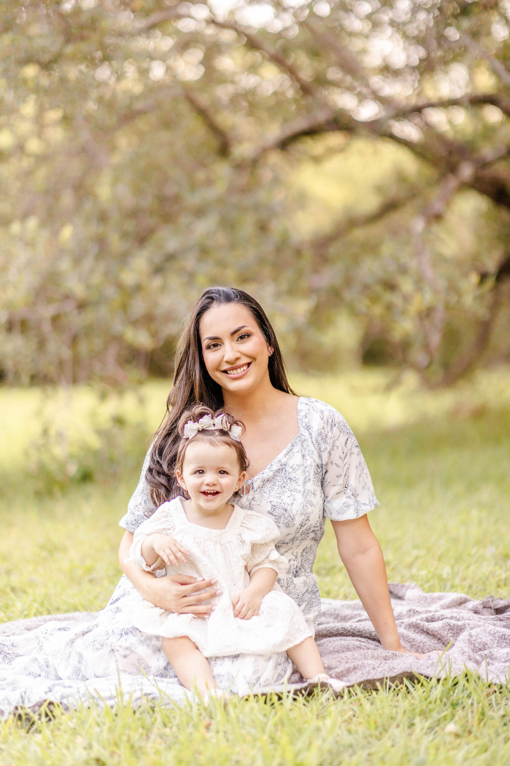 A smiling toddler girl sits in mom's lap in a park lawn at sunset