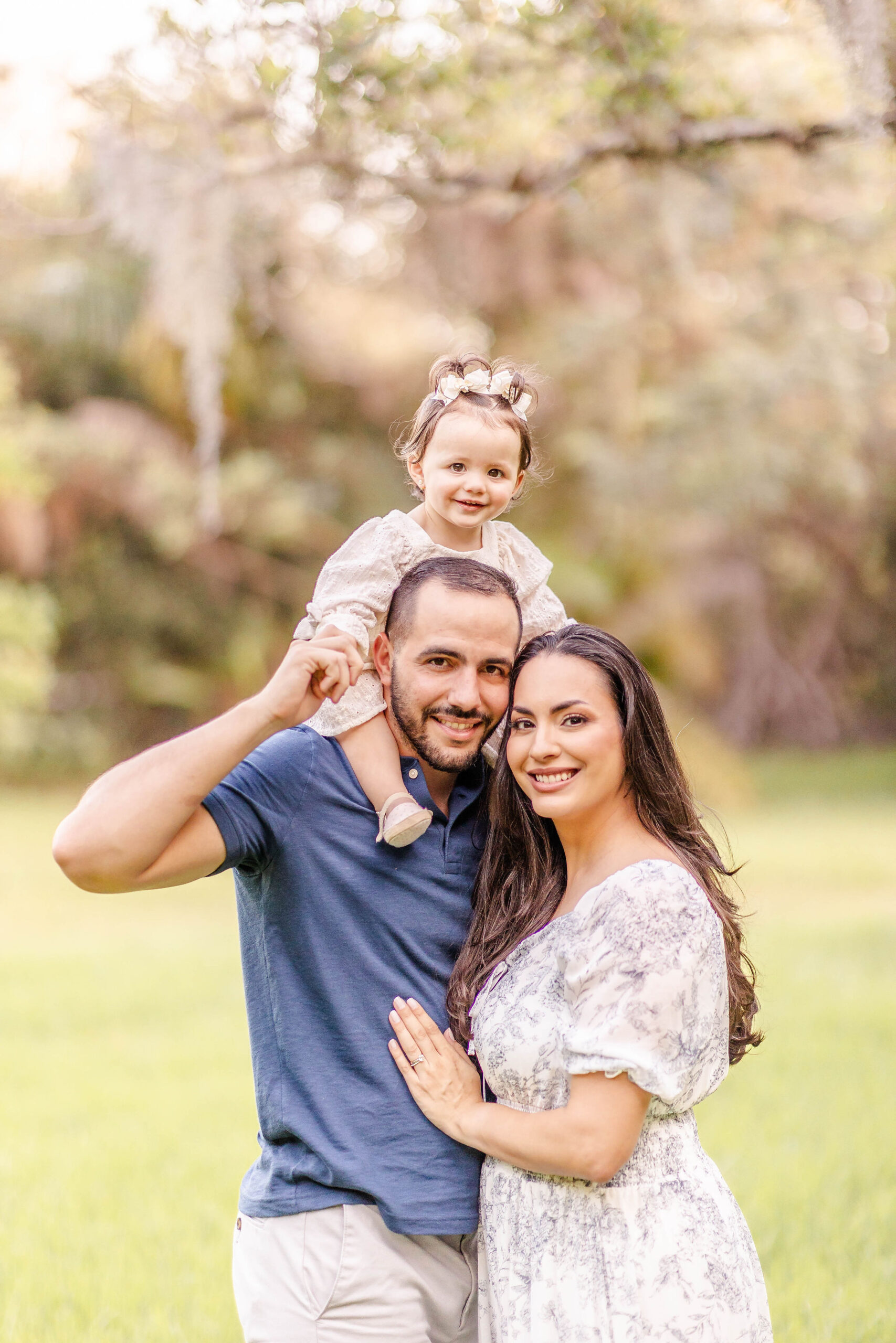 A toddler girl smiles while sitting on dad's shoulders in a park at sunset as mom snuggles with dad before meeting santa in miami
