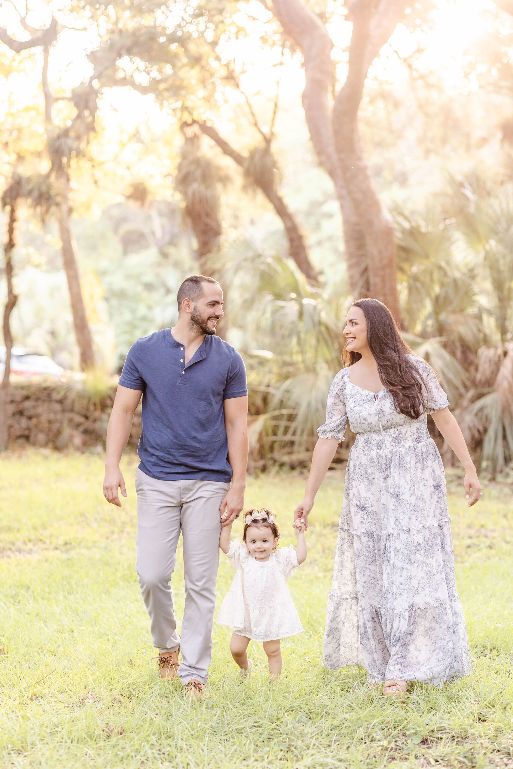 Happy mom and dad in blue walk in a park holding hands with their young toddler daughter at sunset before meeting santa in miami