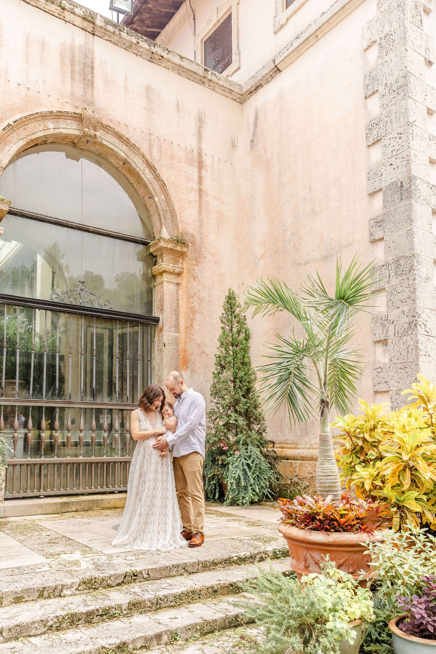 happy mom and dad stand in a garden patio cradling their infant daughter between them