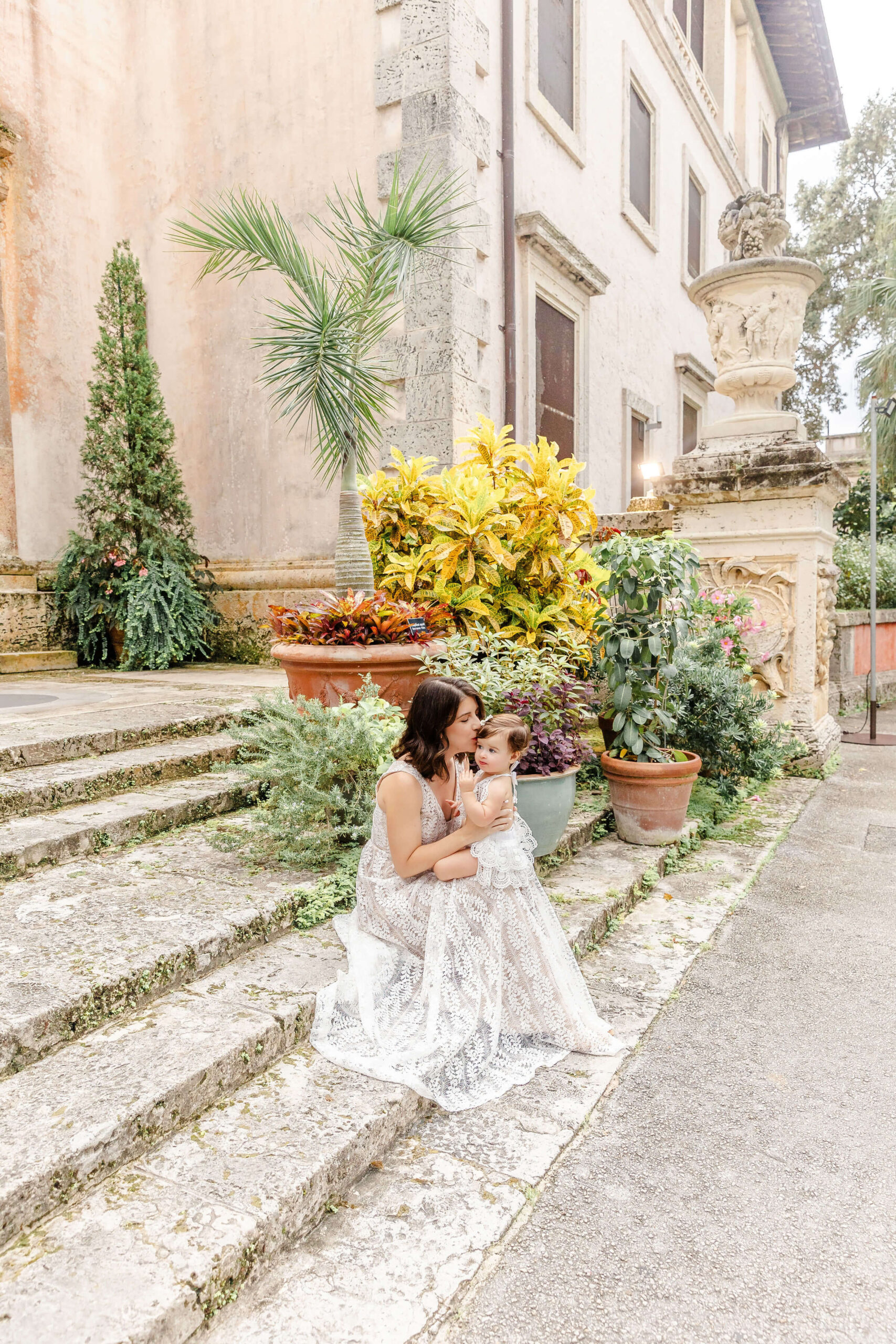 a mom in a white lace dress sits on stone steps kissing her infant in her lap before some christmas events in fort lauderdale