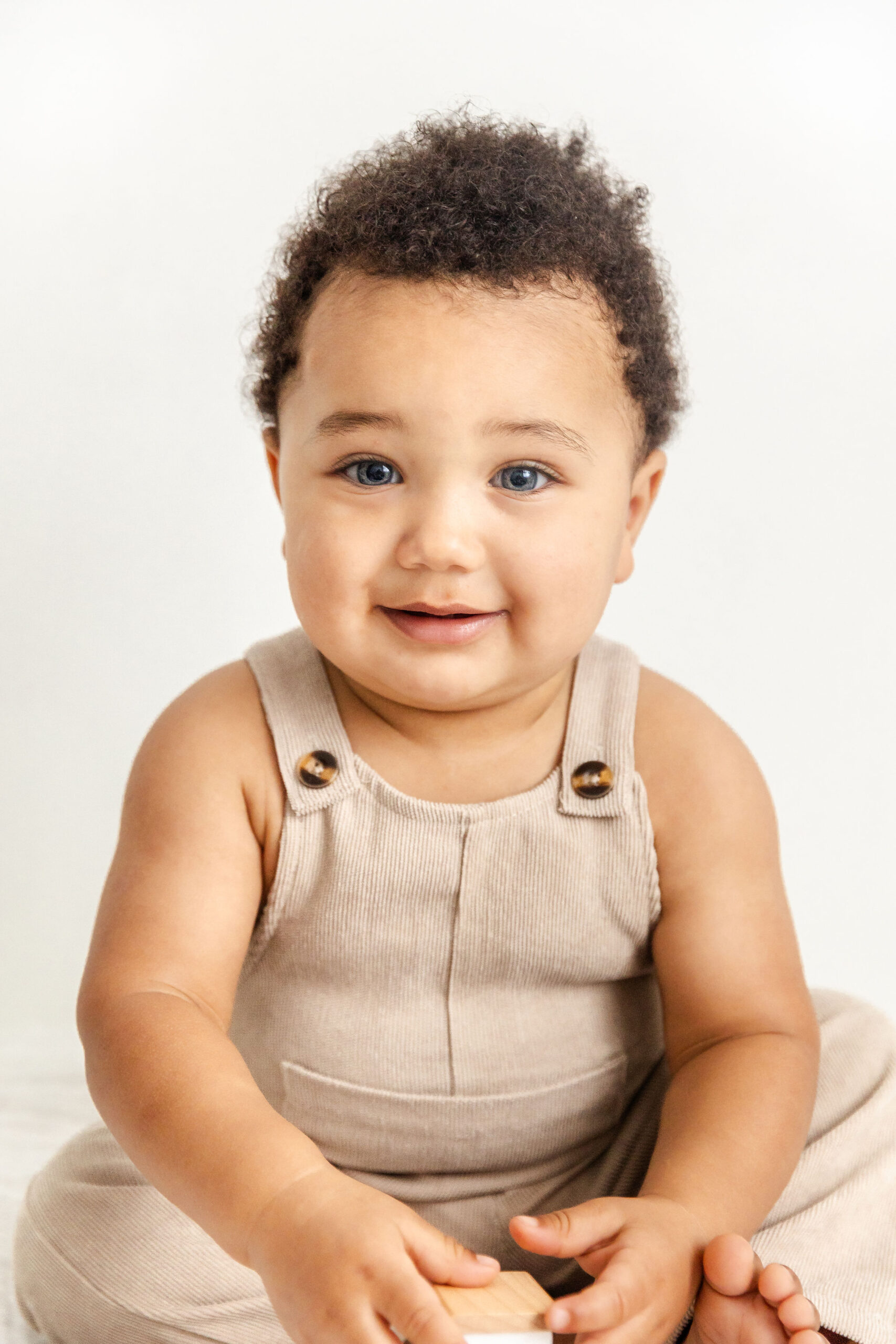 A smiling baby boy in tan overalls sits on the floor of a studio holding a wooden block
