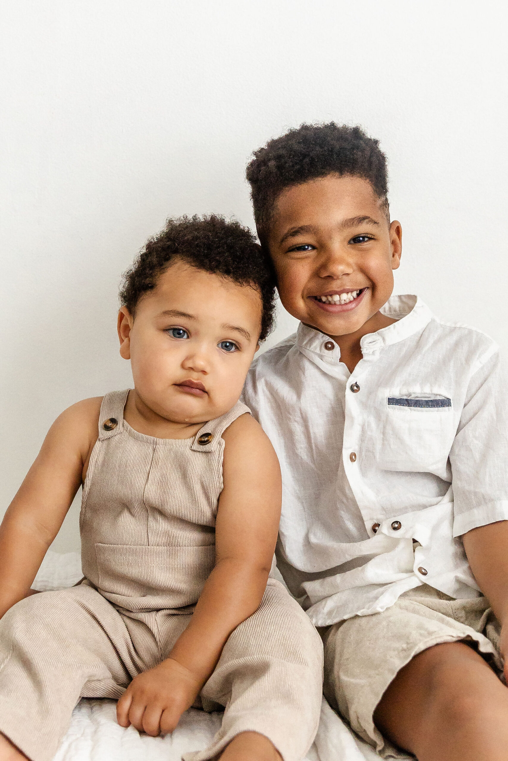 A happy toddler boy in a white shirt sits with his baby brother in a studio on a white bed before visiting fort lauderdale childrens museums