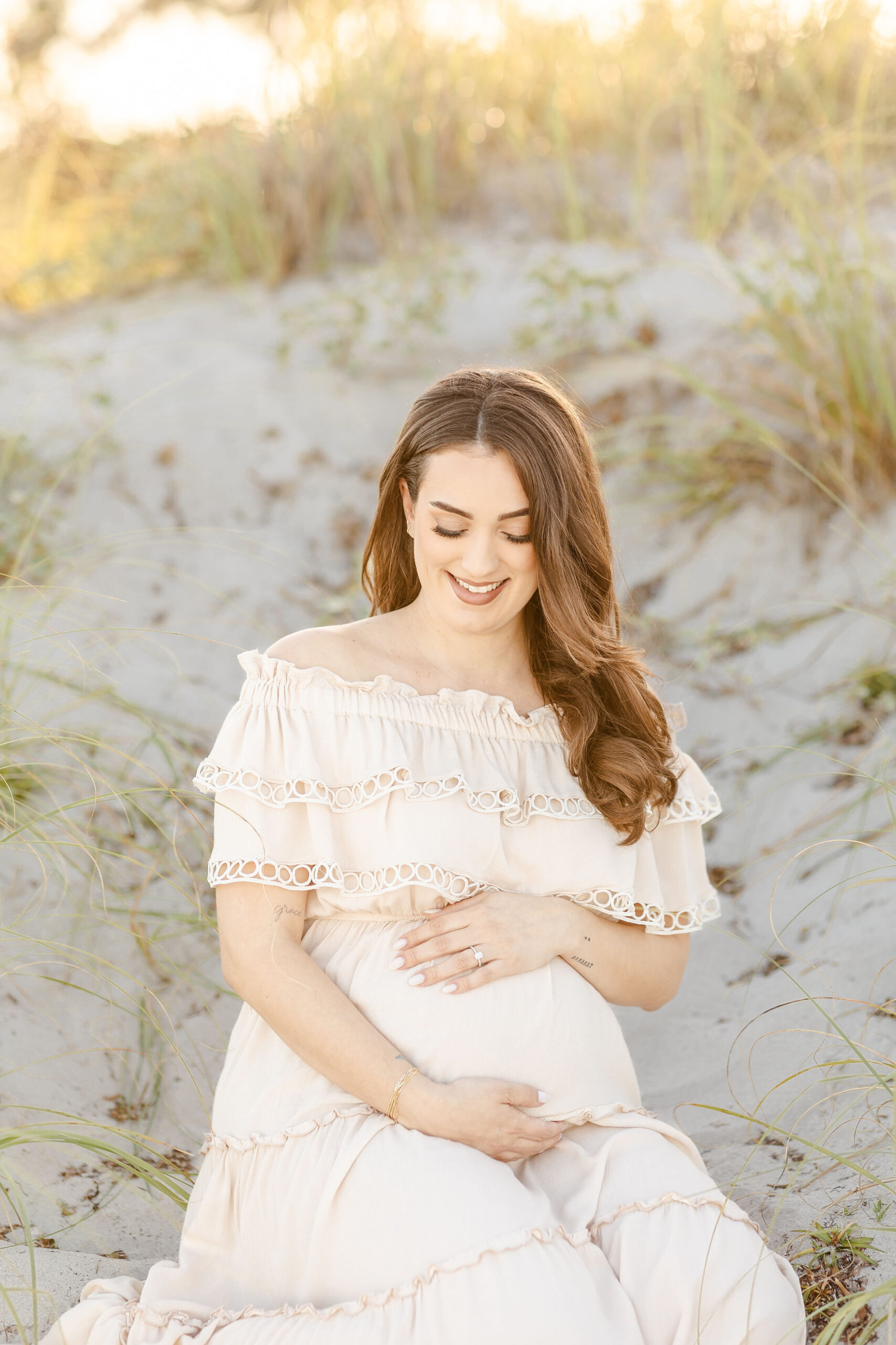A smiling mom to be gazes down to her bump while sitting on a beach dune at sunset after visiting prenatal chiropractors in fort lauderdale