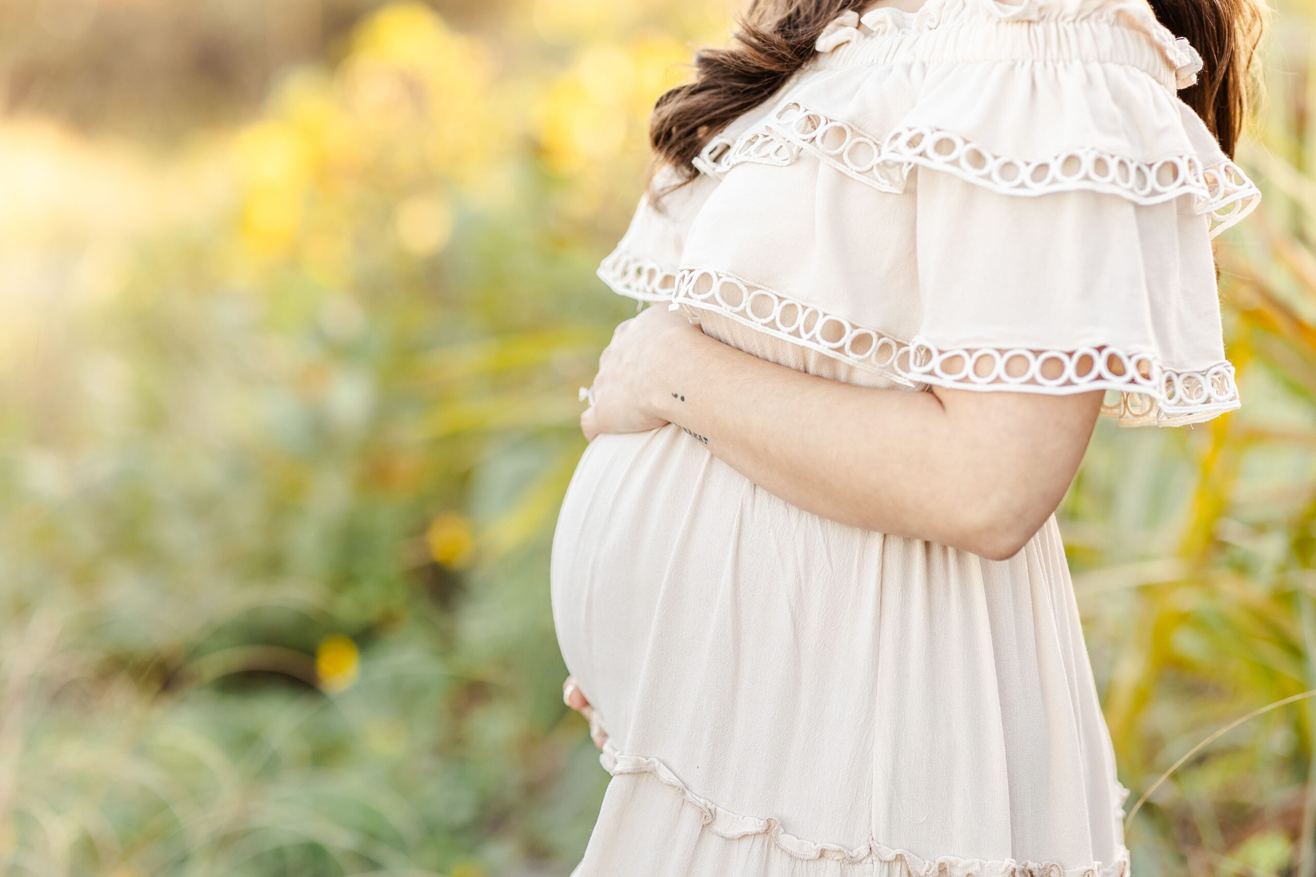 details of the bump of a woman in a cream maternity gown standing in a field with hands on bump