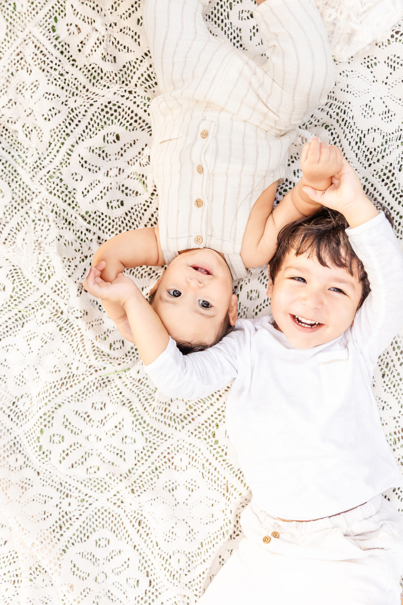Toddler boys laugh while laying on a lace picnic blanket head to head in white outfits