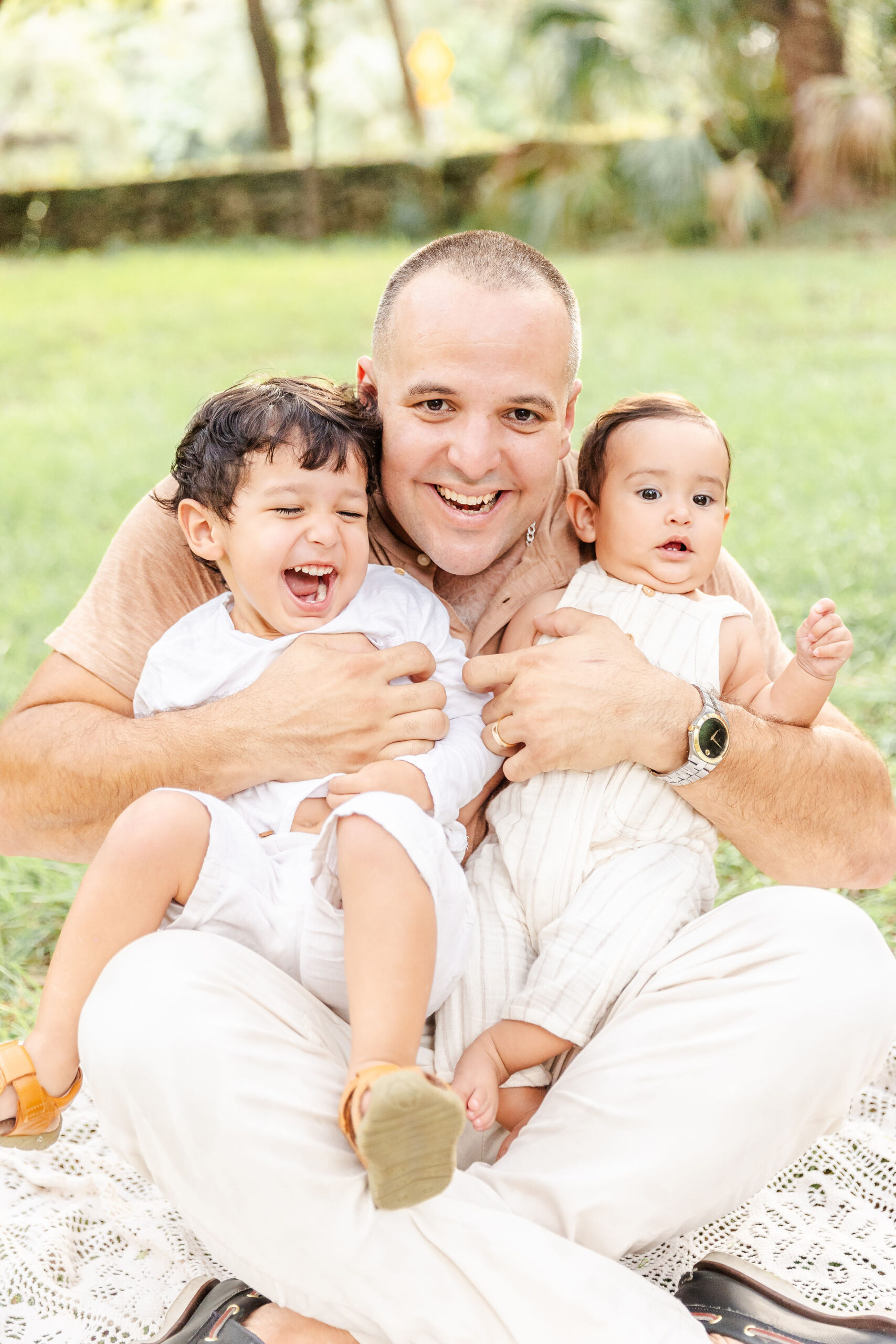 A joyful father sits on a picnic blanket hugging and laughing with his two toddlers thanks to pediatric chiropractors in fort lauderdale