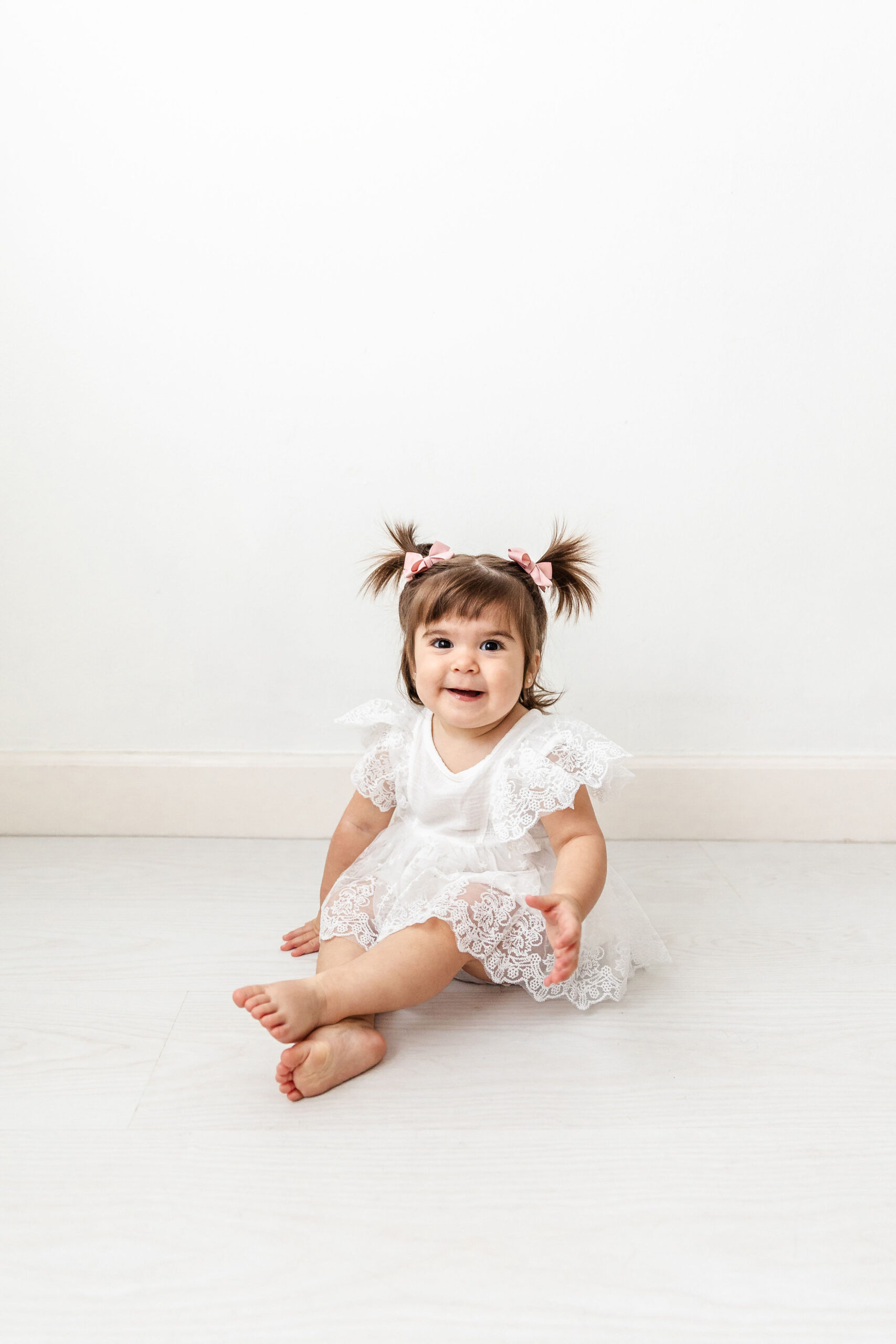 A happy toddler girl in a white lace dress sits on the floor of a studio smiling