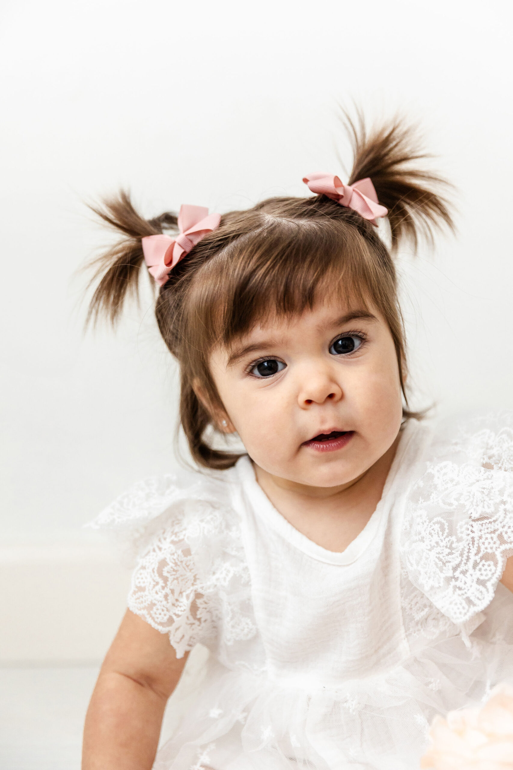 A curious toddler girl with pigtails and a white dress sits in a studio after visiting the tree house playground