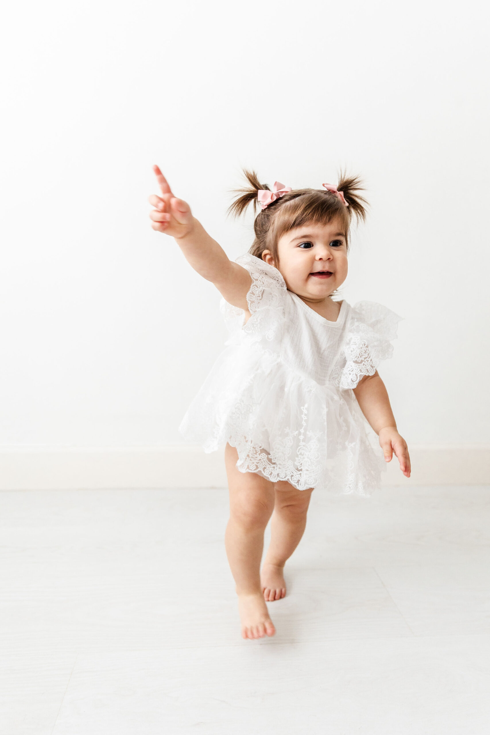 A toddler girl in a white dress plays in a room with a finger in the air before visiting the tree house playground