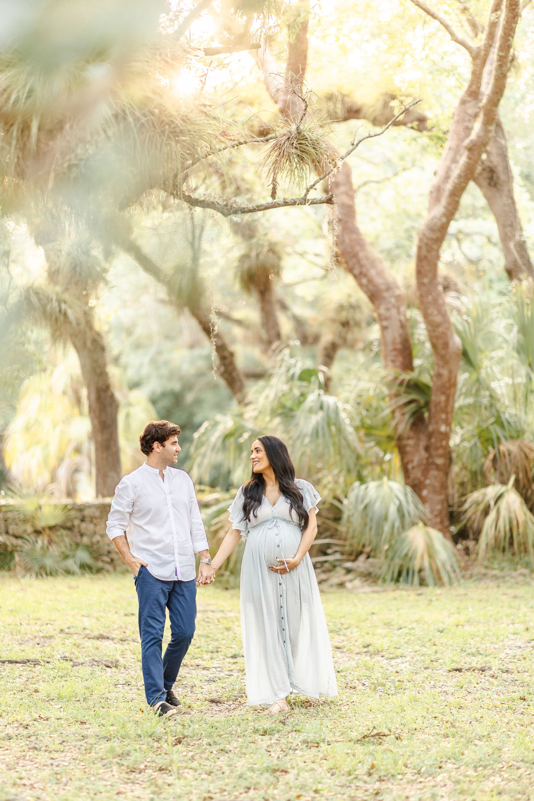 A mom to be walks with a hand under her bump and holding hands with her husband in a white shirt in a florida park at sunet