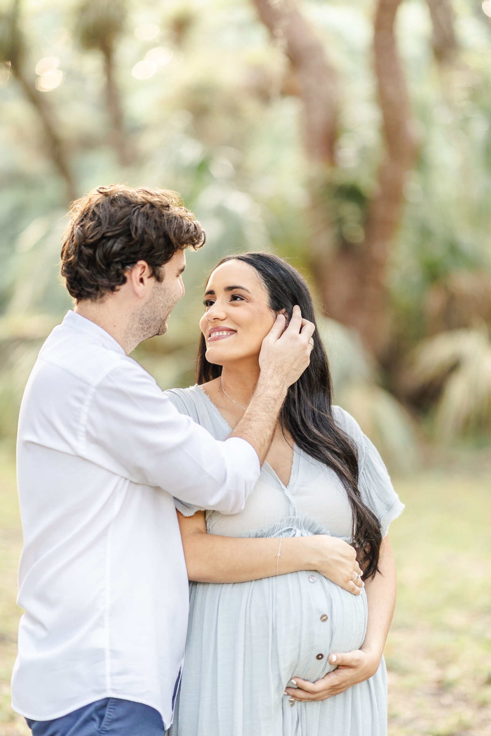 A man in a white shirt pulls the hair behind his pregnant wife's ear while they stand in a park lawn at sunset after visiting Wisdombirth