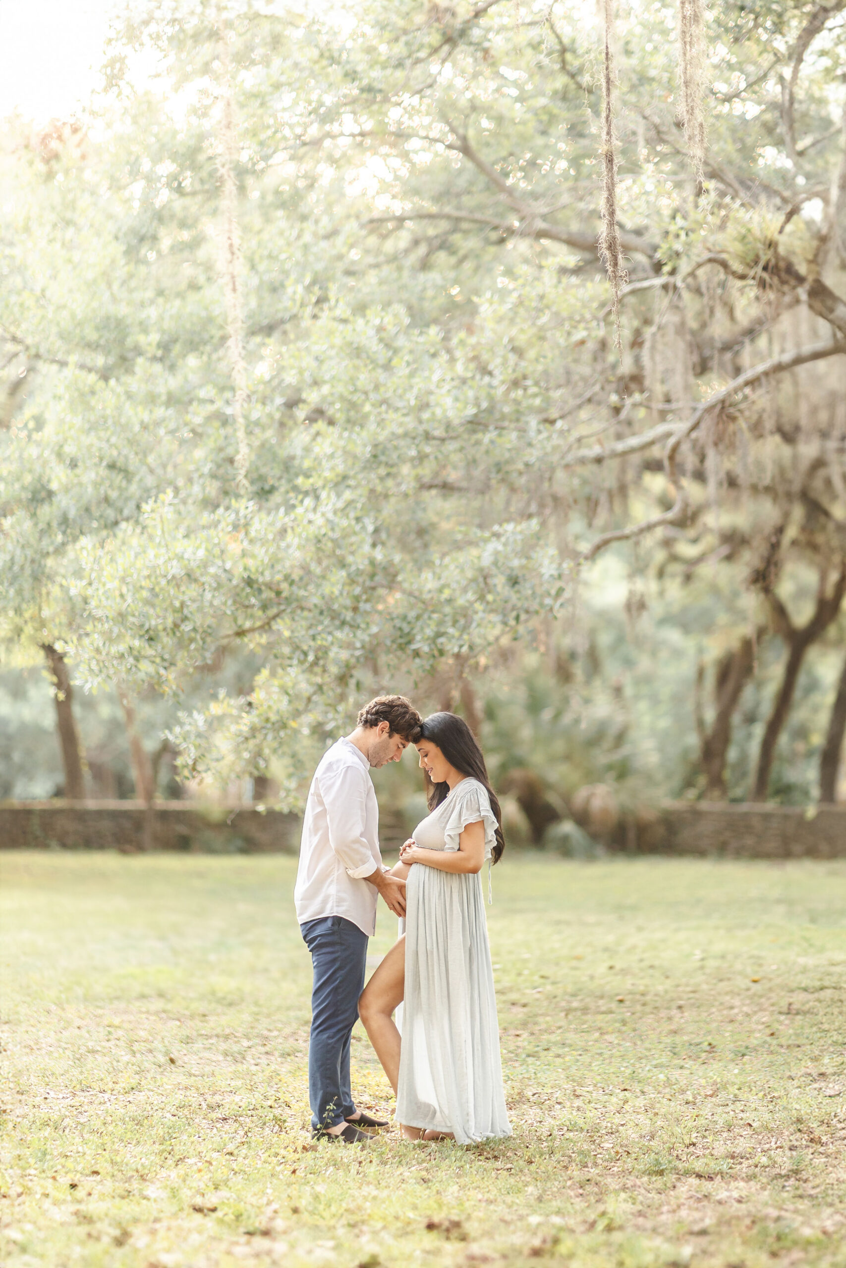 Expecting parents stand in a park with hands on the bump at sunset in a white maternity gown after visiting Wisdombirth