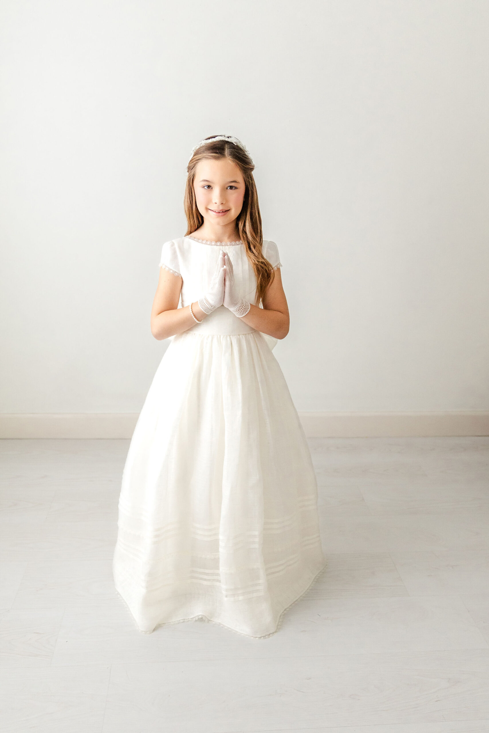 A young girl holds her hands in prayer while smiling in a white gown in a studio