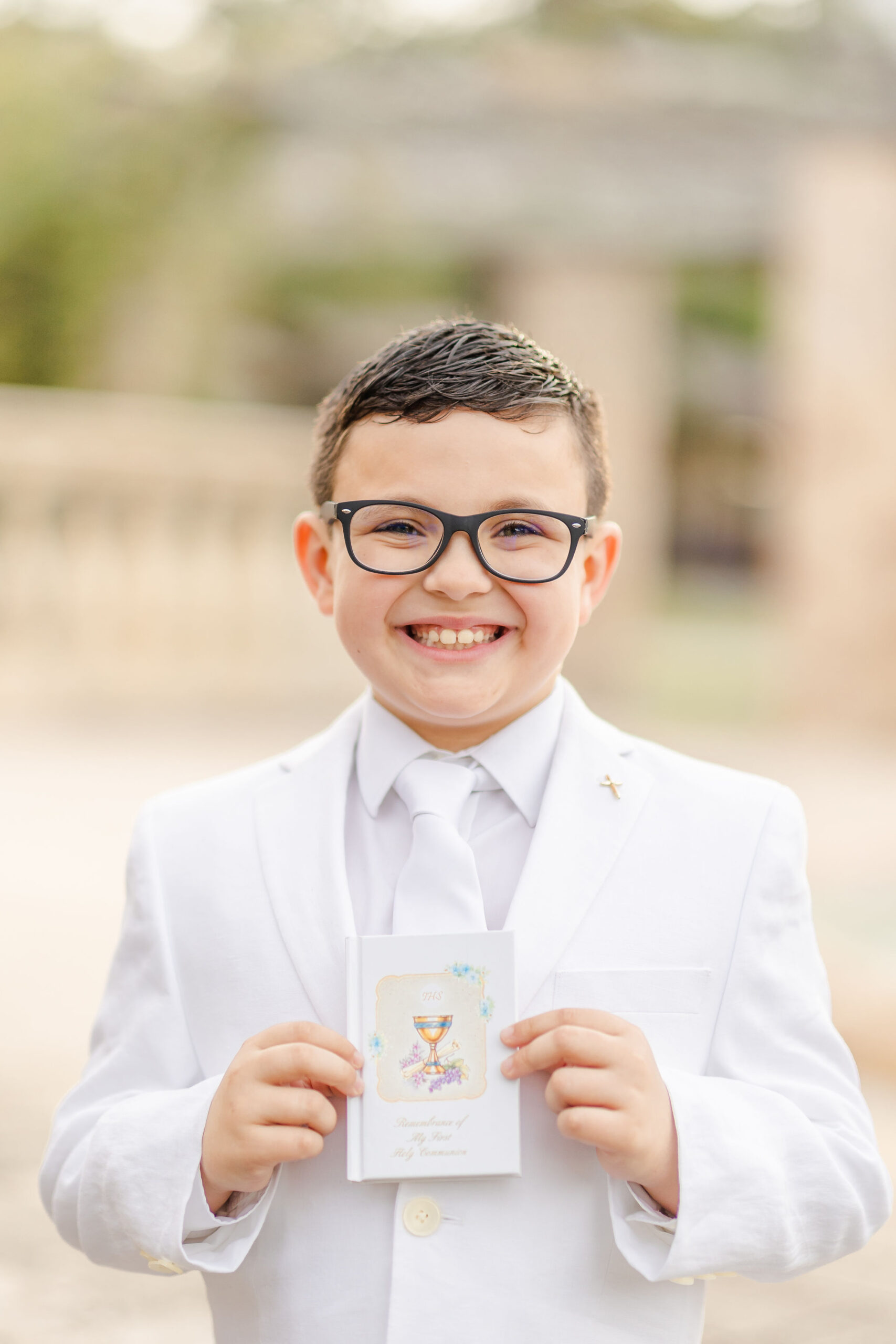 A smiling young boy with glasses and an all white suit holds his prayer book and smiles after finding great first communion suits in miami