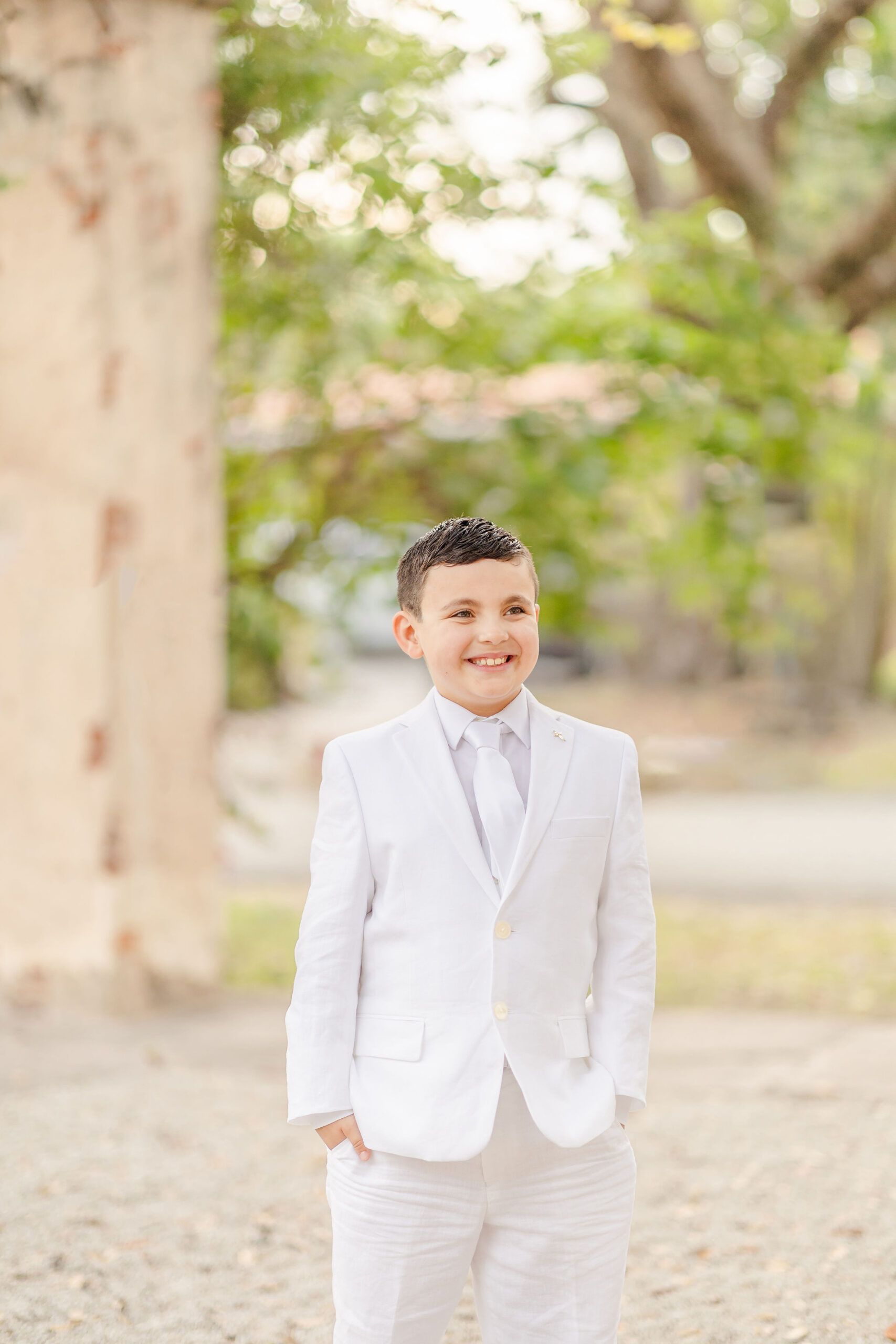 A happy young boy stands in his white first communion suits in miami with hands in his pockets outside under trees