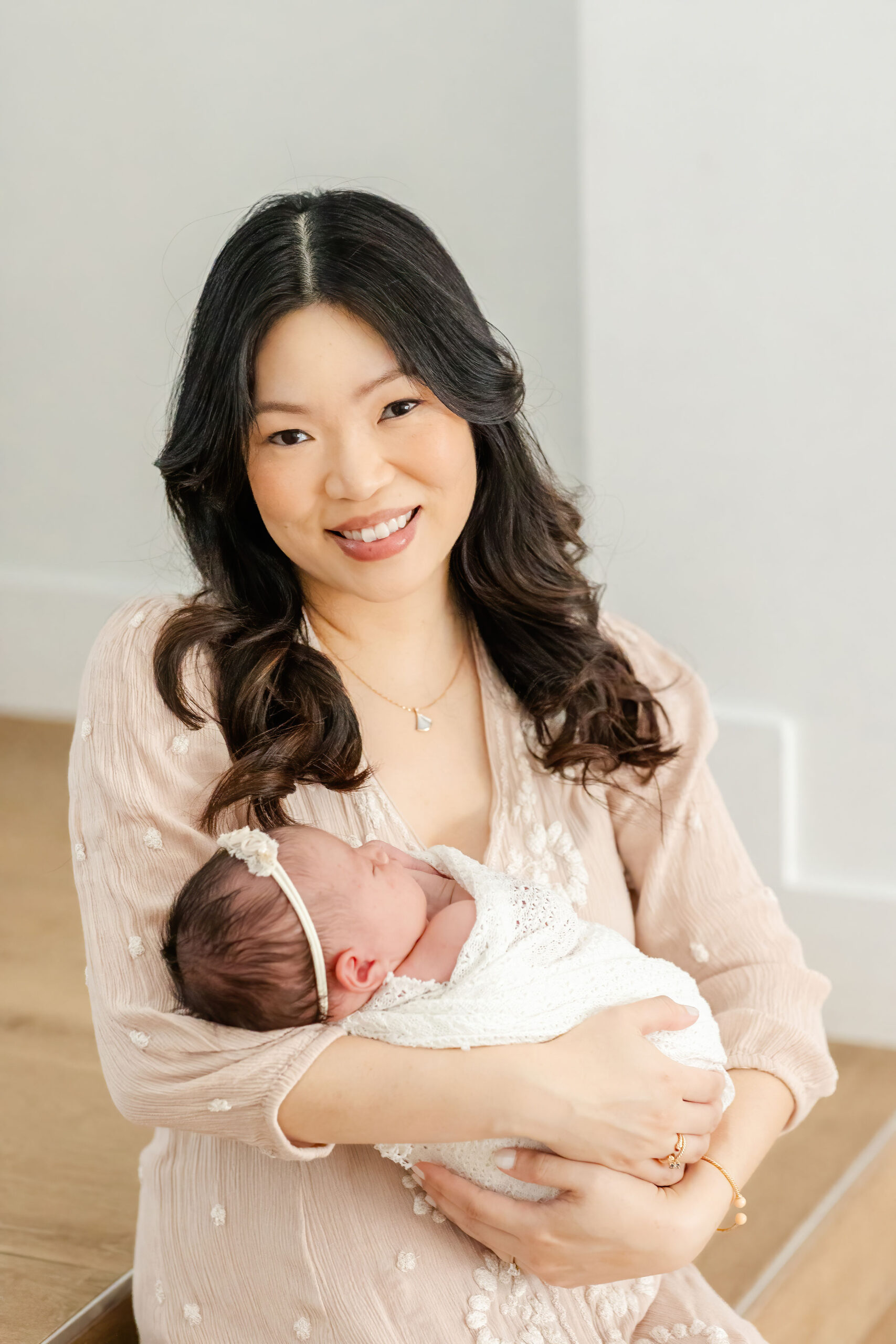 A smiling mother in a cream dress holds her sleeping newborn daughter while sitting on wood steps after finding Baby stores in coral gables