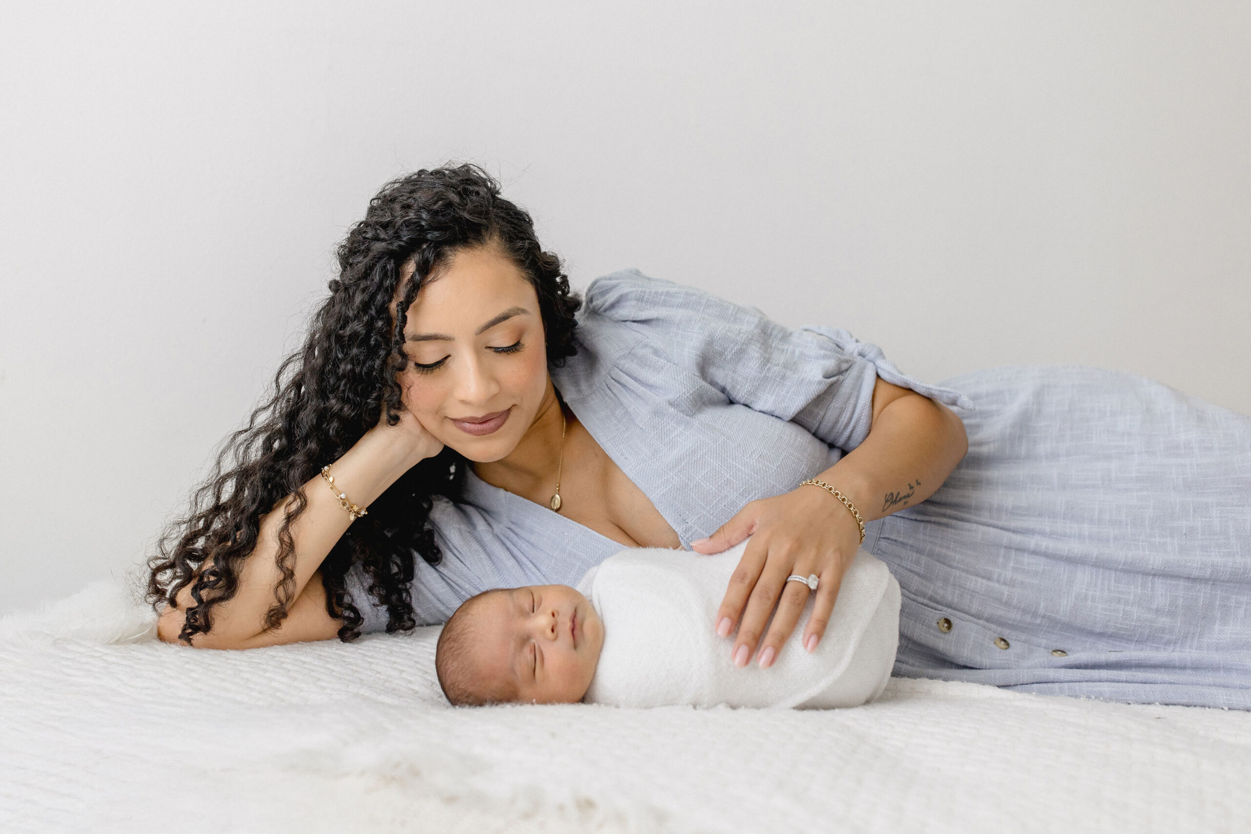 A happy new mom in a blue dress lays alongside her sleeping newborn on a bed