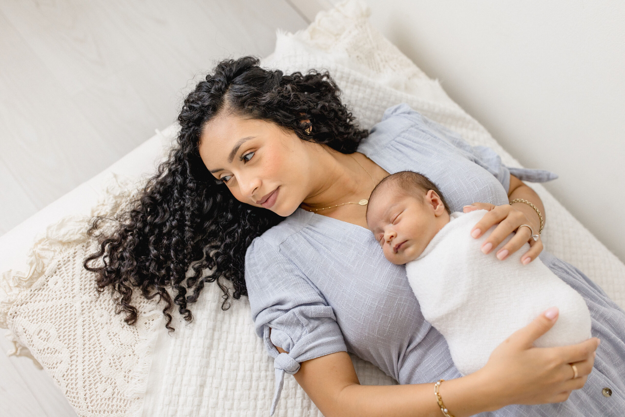 A happy mother in a blue dress lays on a bed in a studio with her newborn sleeping on her stomach after meeting lactation consultants in coral gables