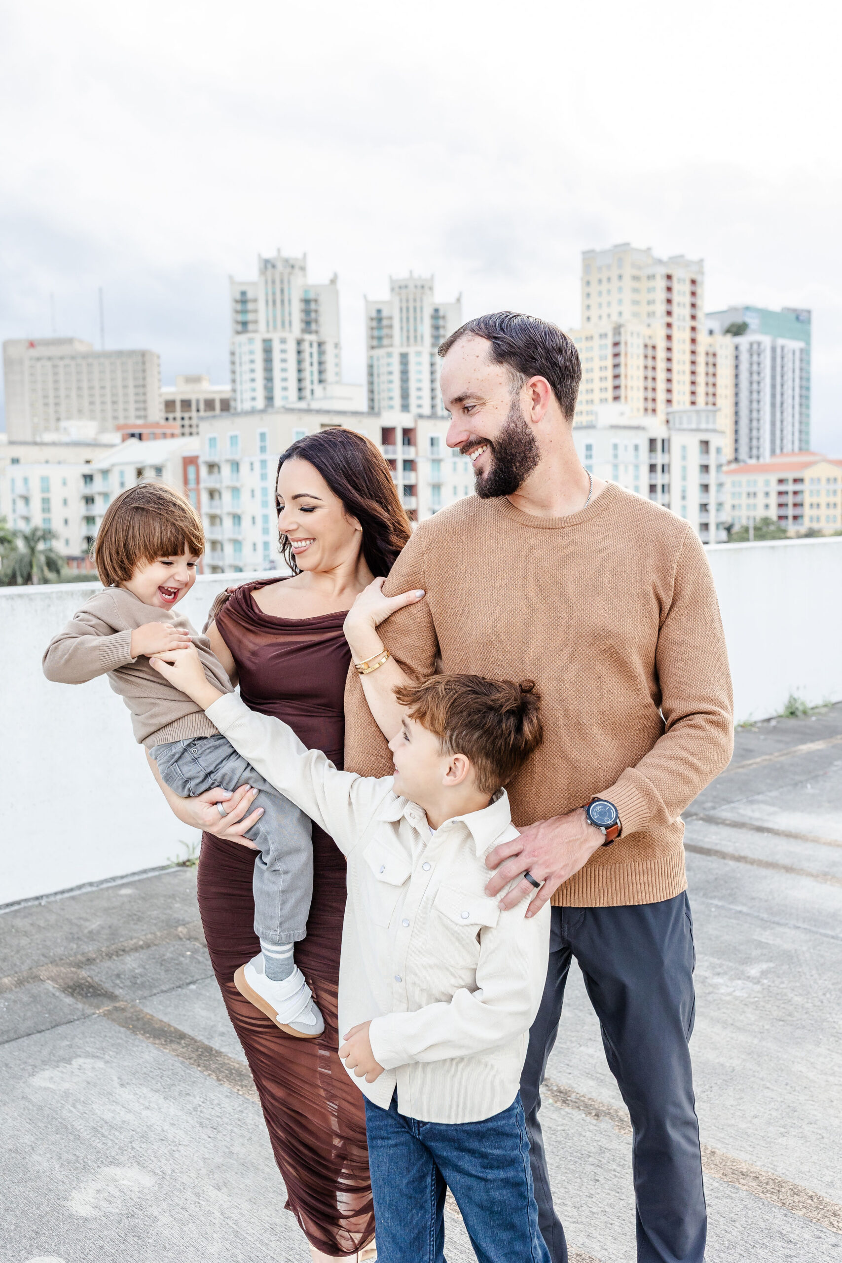 A mom and dad smile in brown while playing with their toddler sons on a cement dock