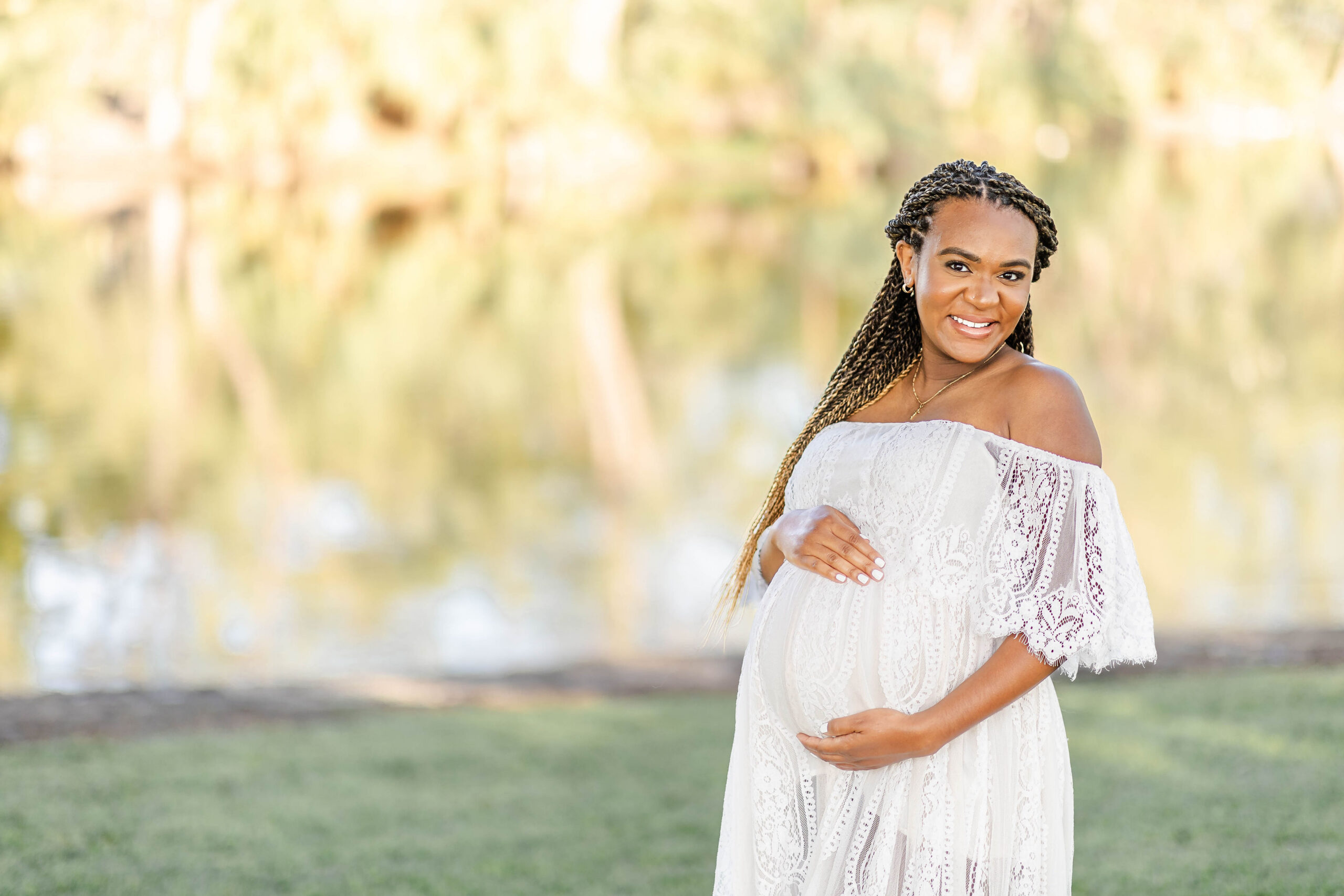 A mother to be in a lace maternity gown smiles with hands around her bump in a park