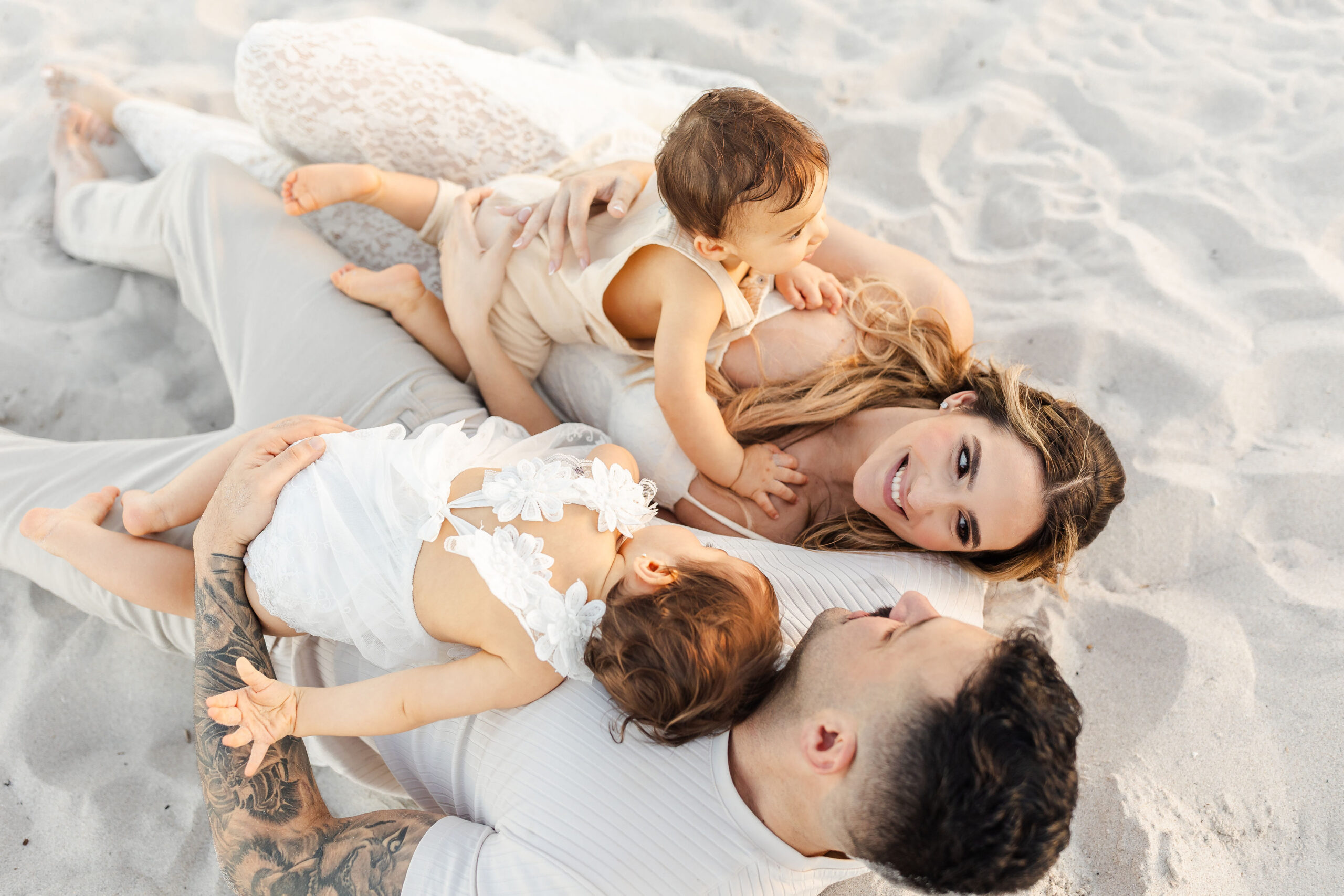 A look down at a mom and dad laying in the sand with their toddler son and daughter laying on them