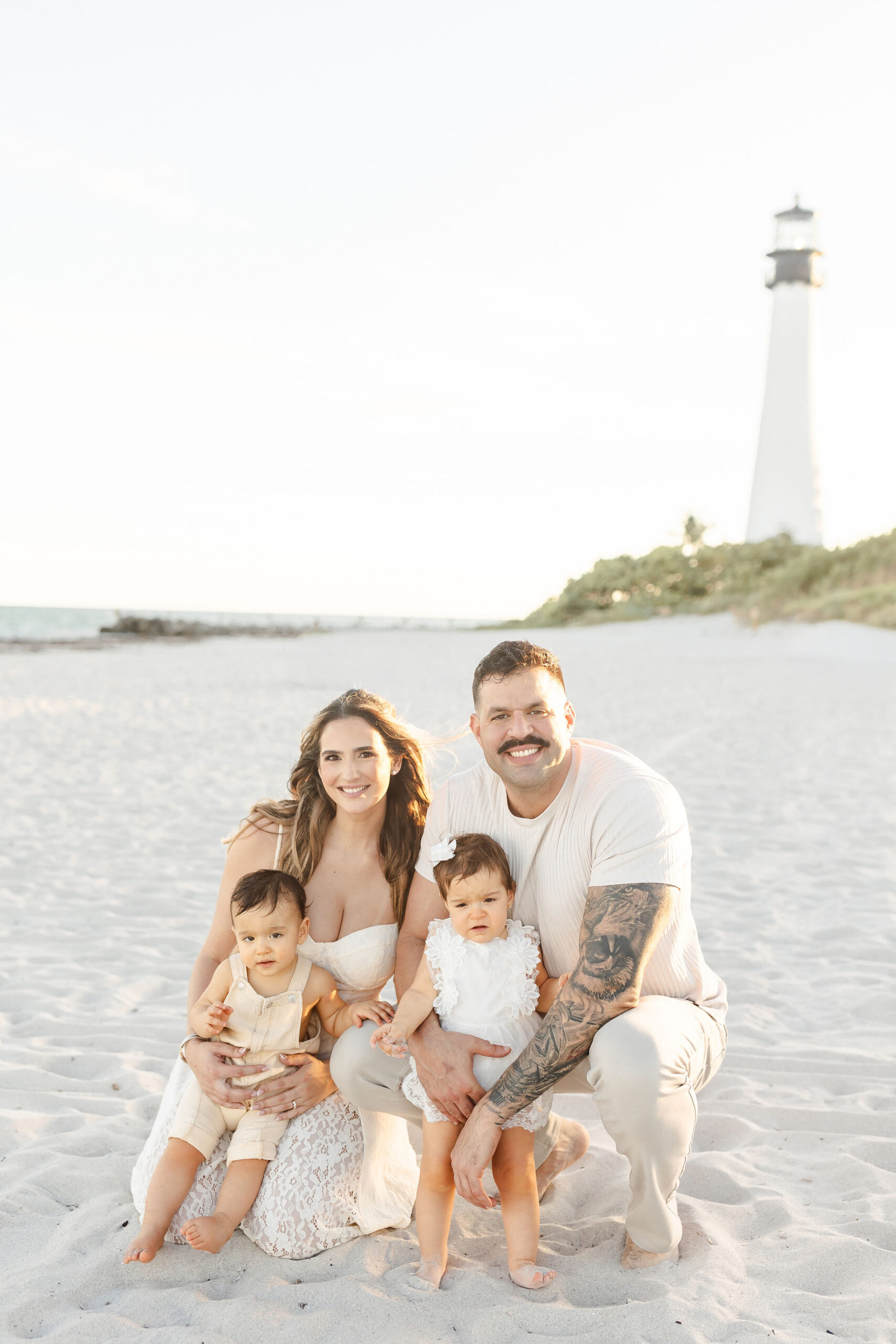 A mom and dad in white kneel and hug their toddler son and daughter on a beach at sunset after visiting daycares in coral gables
