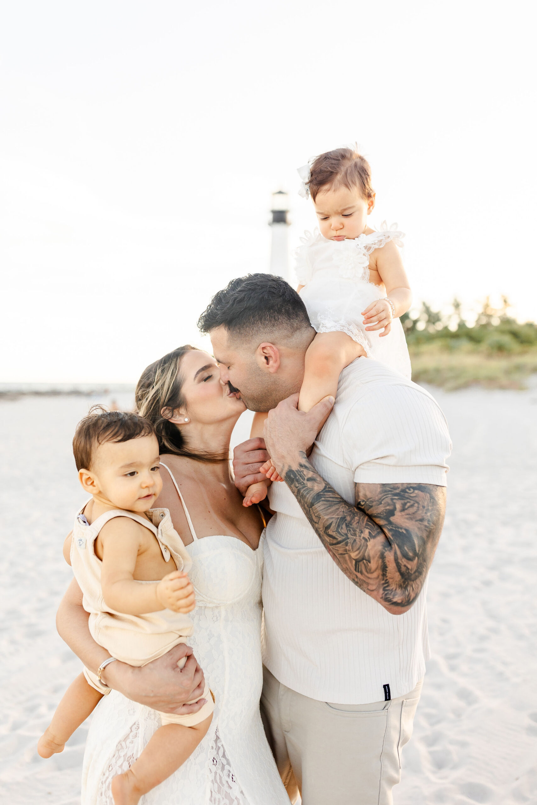 Mom and dad kiss while holding their toddler son and daughers on a beach after finding daycares in coral gables