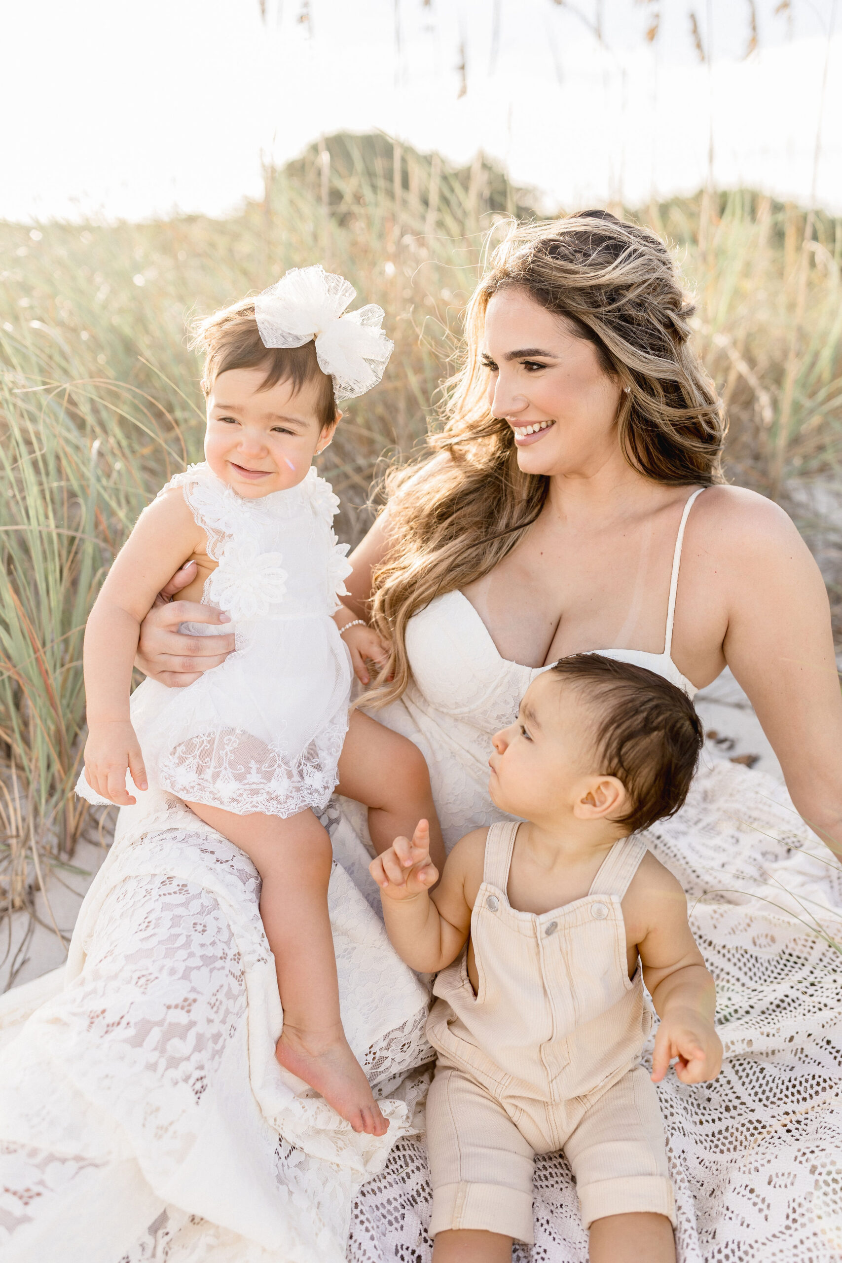 A smiling mother in a white lace gown sits on a blanket in the beach with her toddler son and daughter