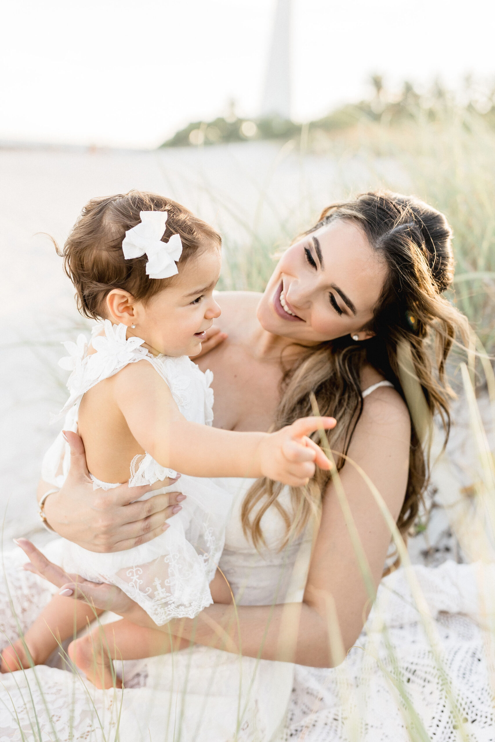 A smiling mother in a white dress plays with her toddler daughter also in white lace in a beach dune at sunset after some mommy and me classes in coral gables