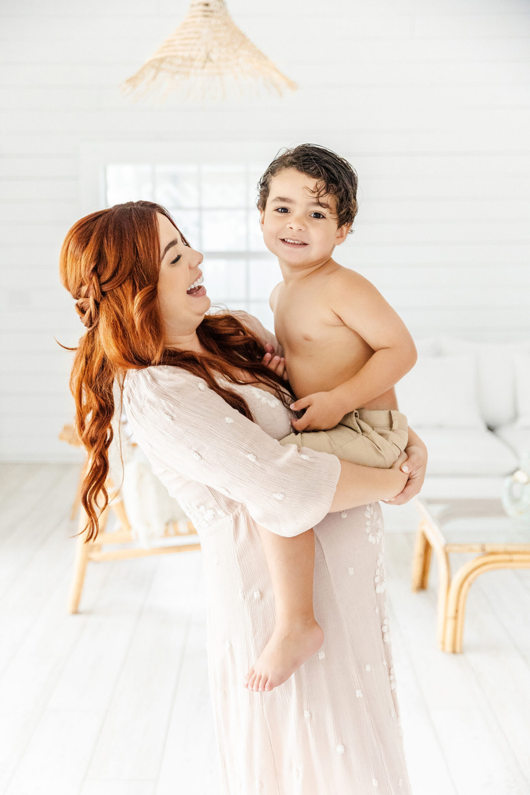 A laughing mother in a white dress holds her toddler son on her pregnant bump in a studio after some pelvic floor therapy in coral gables