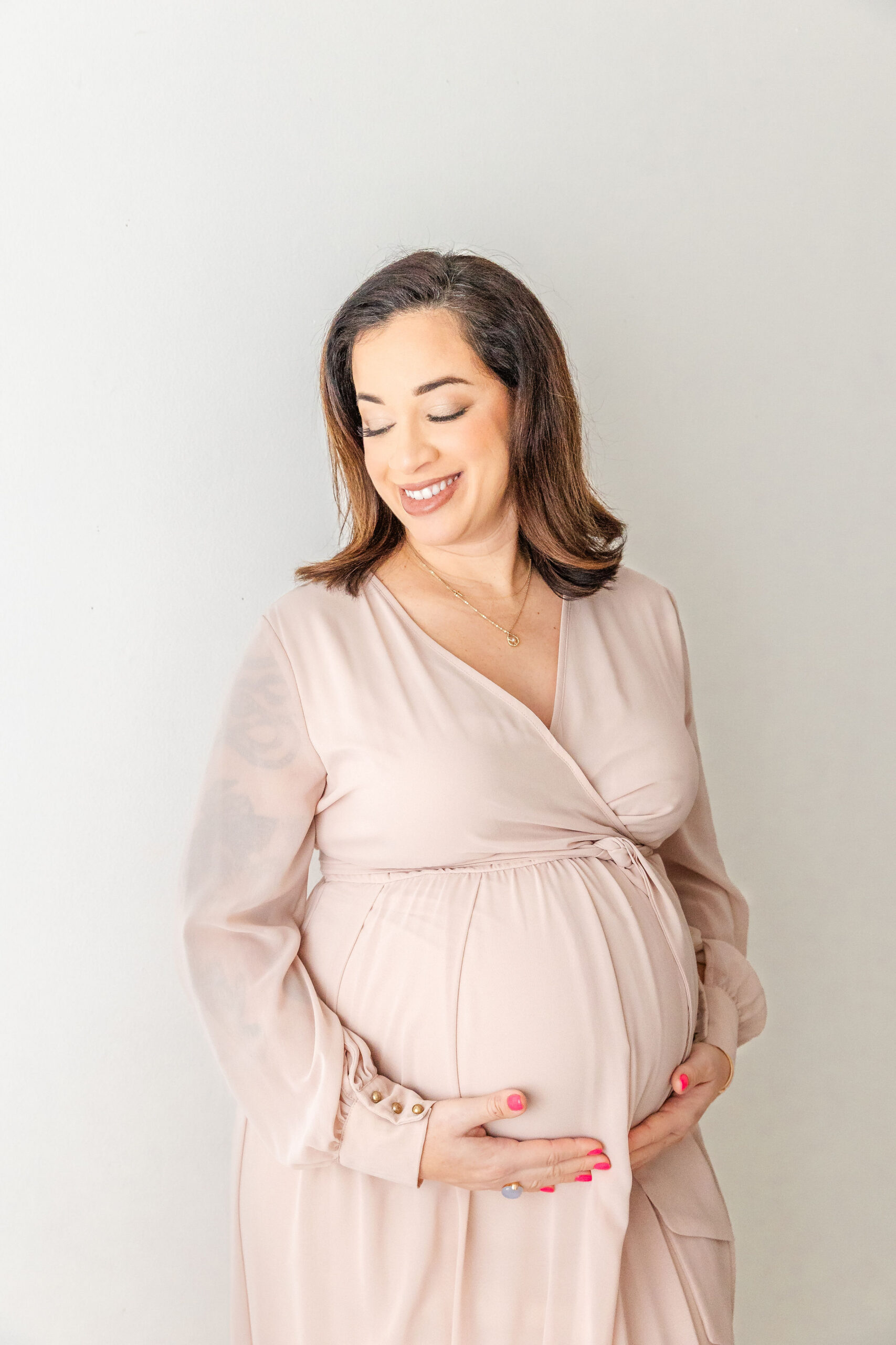 A happy mother to be smiles down her shoulder while holding her bump in a studio in a pink dress after enjoying prenatal fitness in coral gables