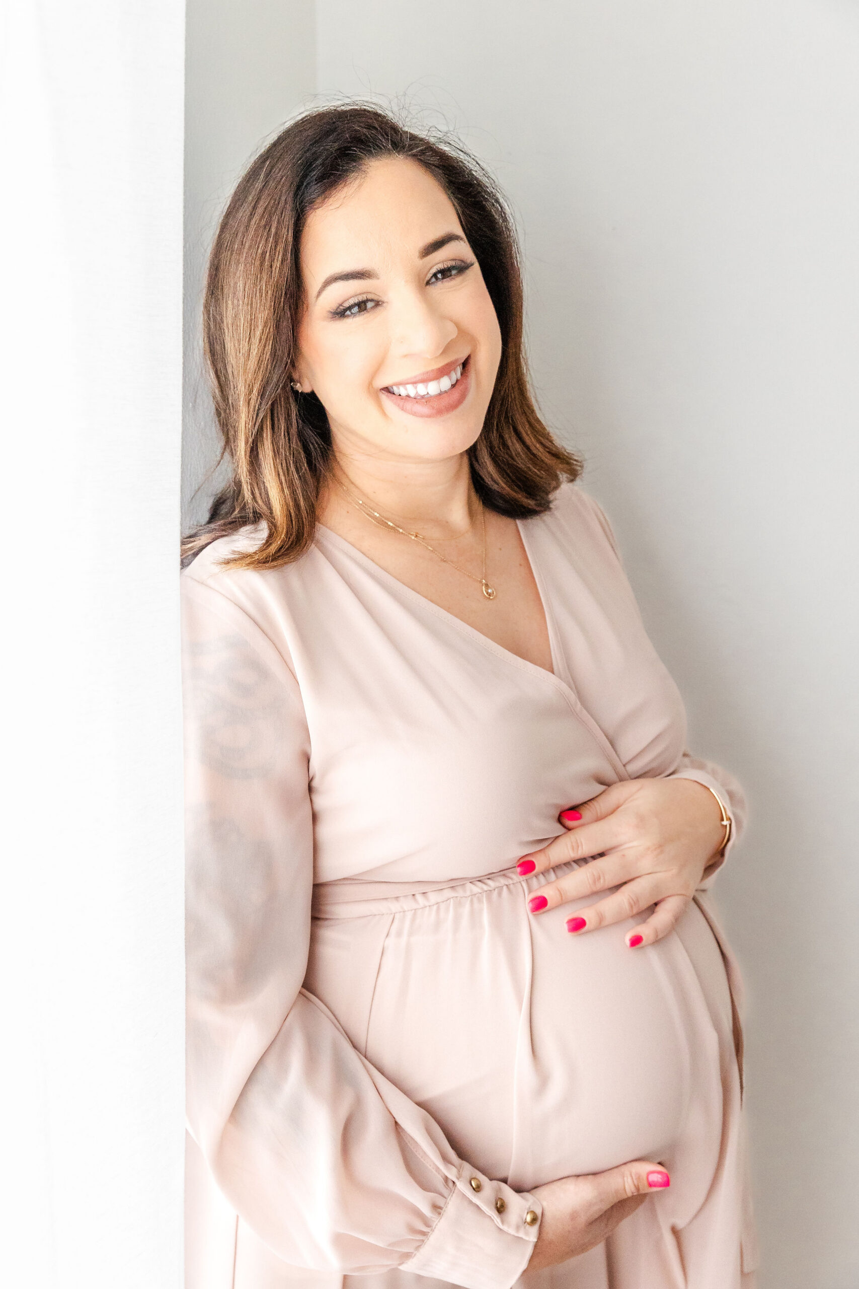 A happy pregnant woman smiles while standing in a pink maternity gown in a studio after some prenatal fitness in coral gables