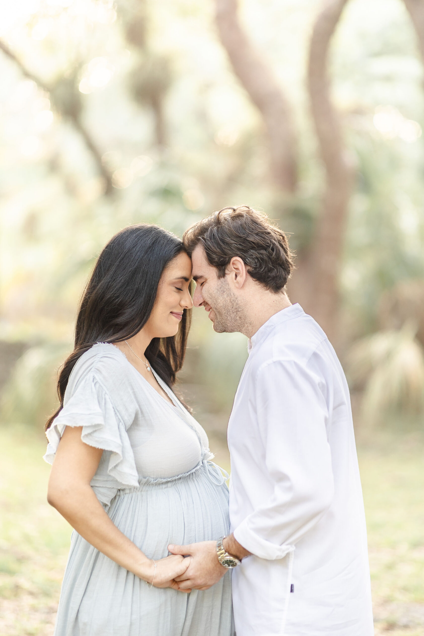 A happy expecting couple hold hands and touch foreheads while smiling in a park at sunset