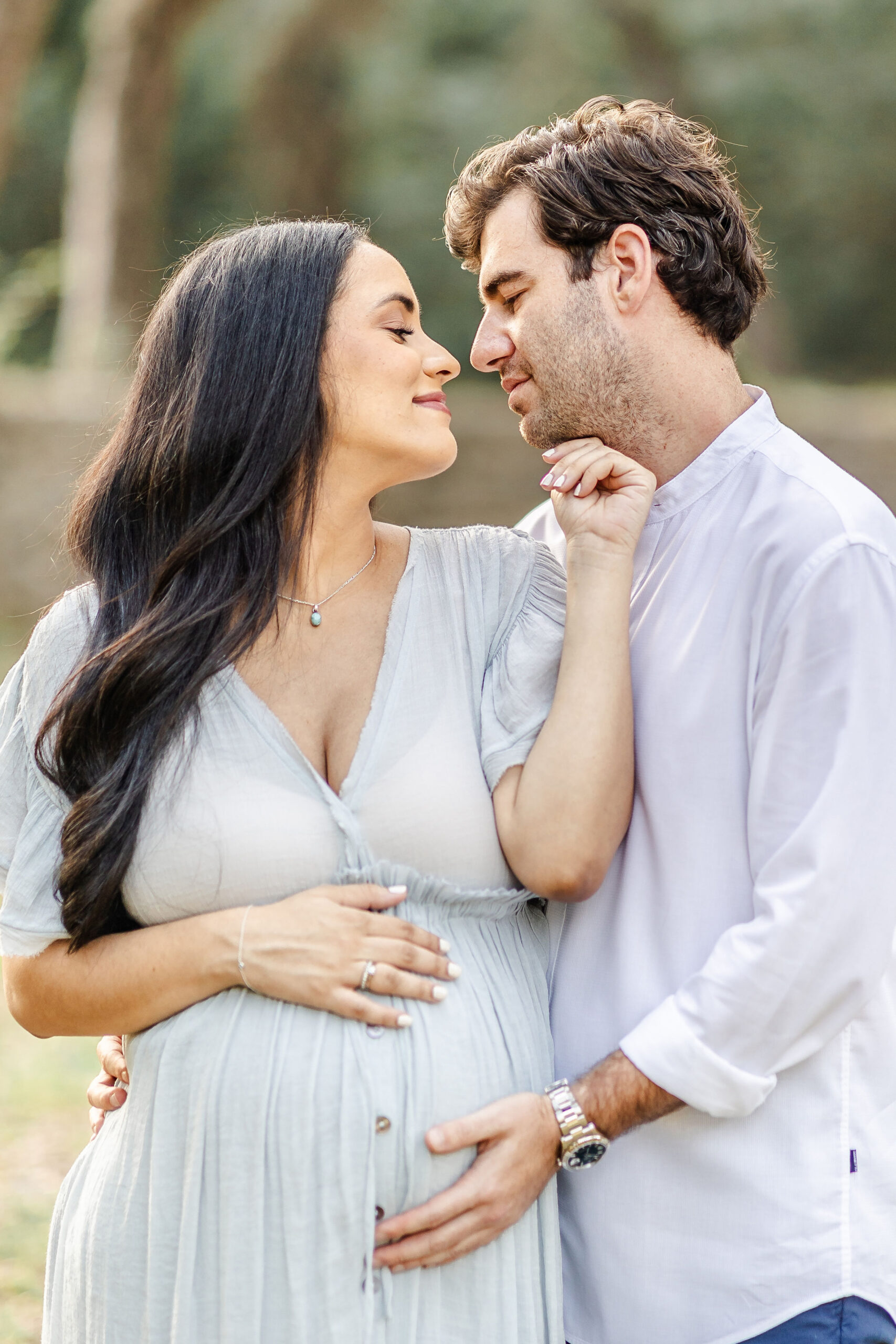 A happy pregnant couple snuggles and leans in for a kiss with hands on the bump in a park after enjoying baby shower venues in coral gables